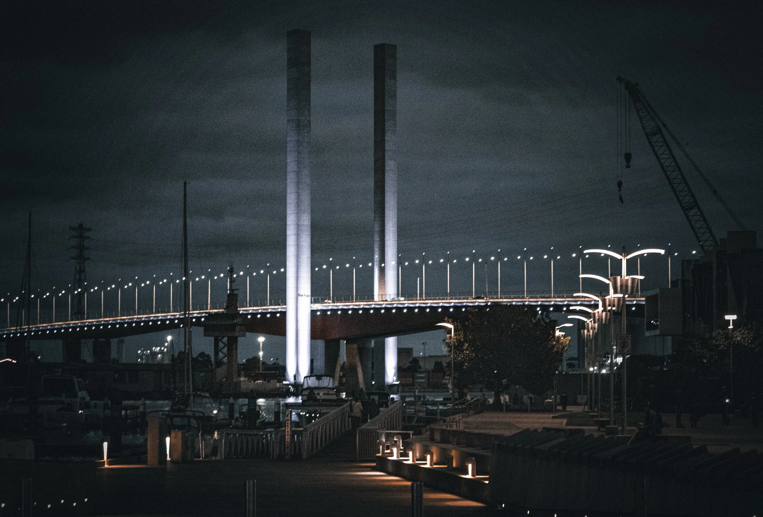 Nighttime view of a lit-up bridge with two large, illuminated vertical supports and a crane nearby.