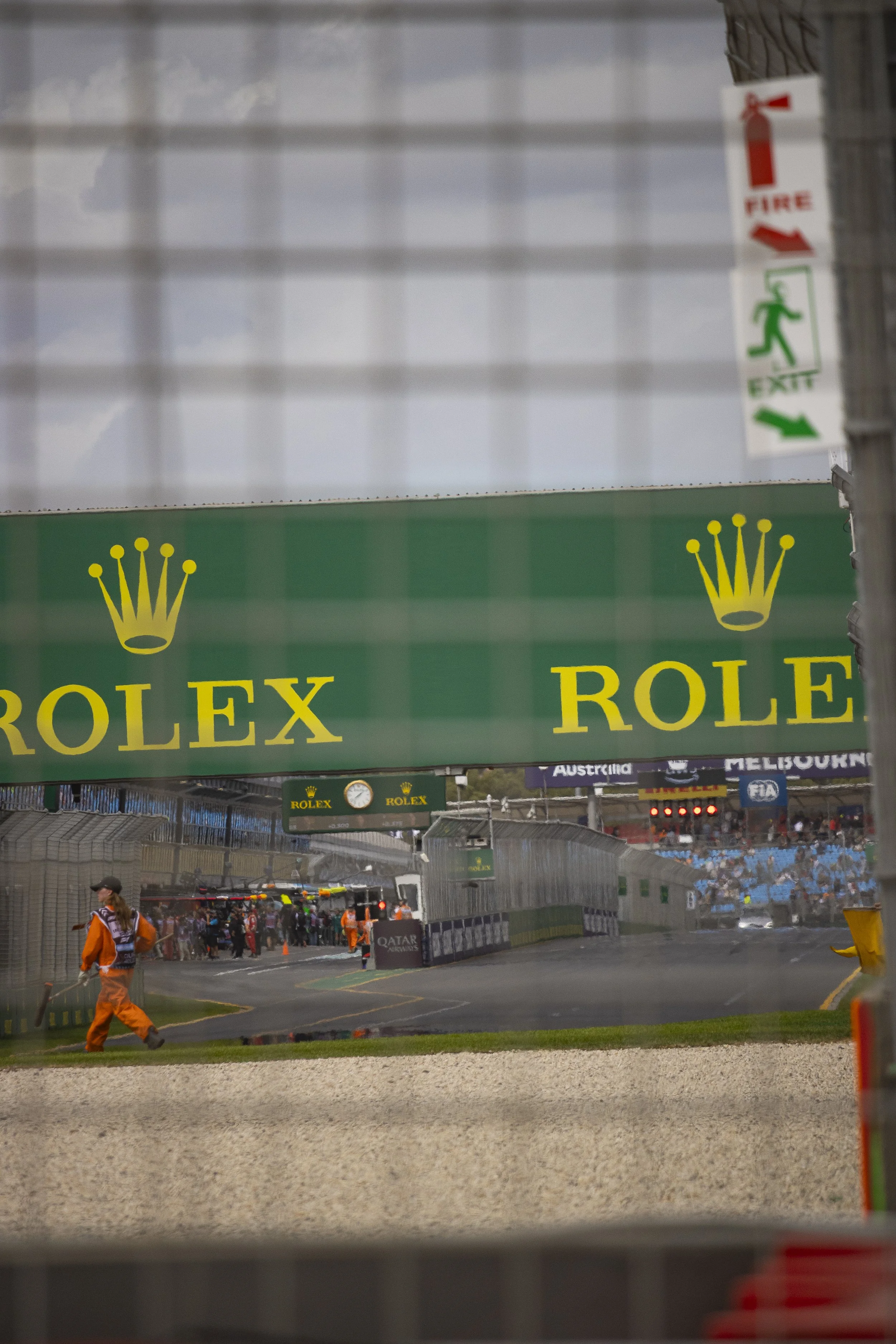 An image taken at a motor racing event showing a section of the racetrack, with the exit signs, race officials, and spectators in the background. Large green Rolex banners are visible.