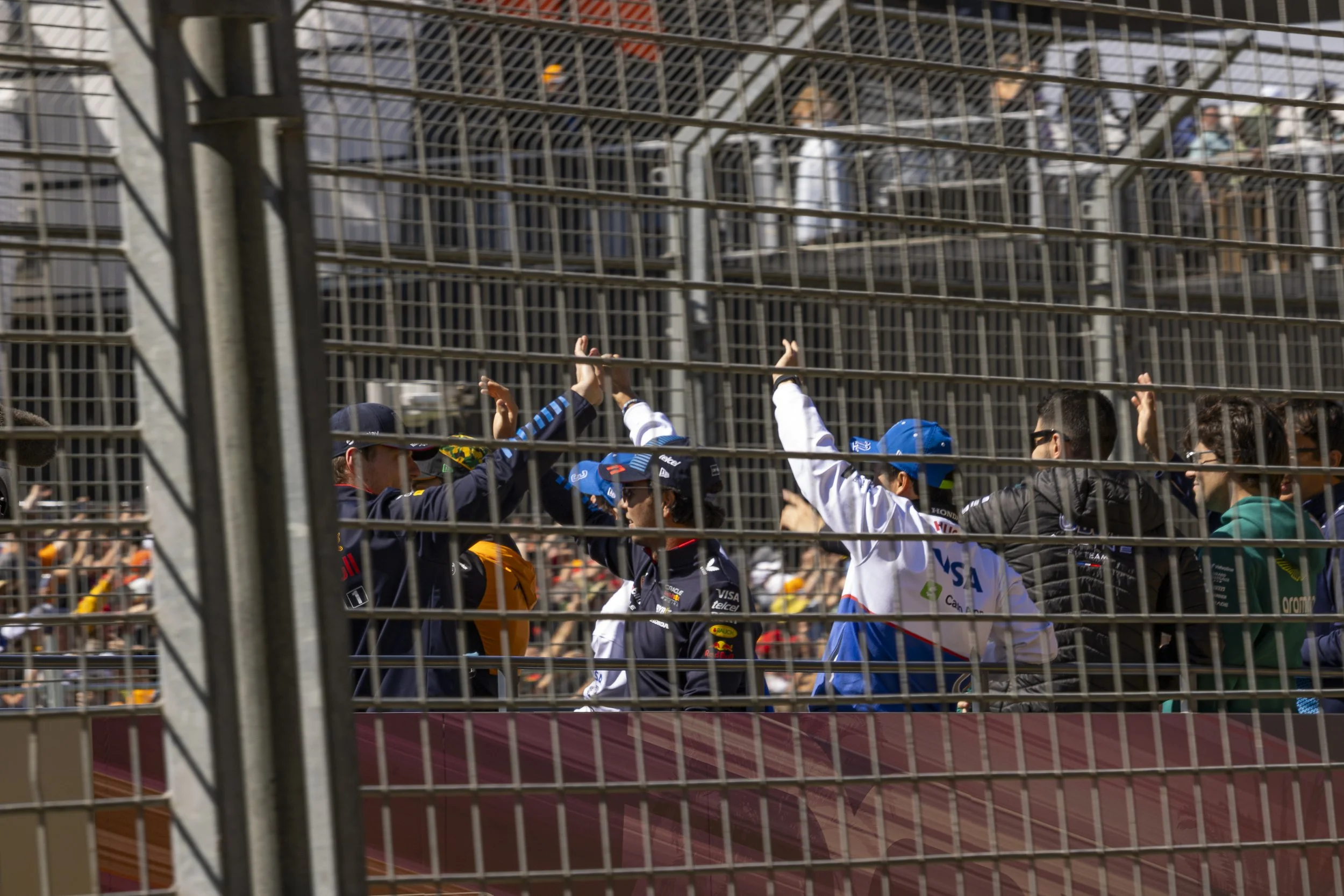 Formula 1 drivers and team members high-fiving behind a metal fence at a race track.