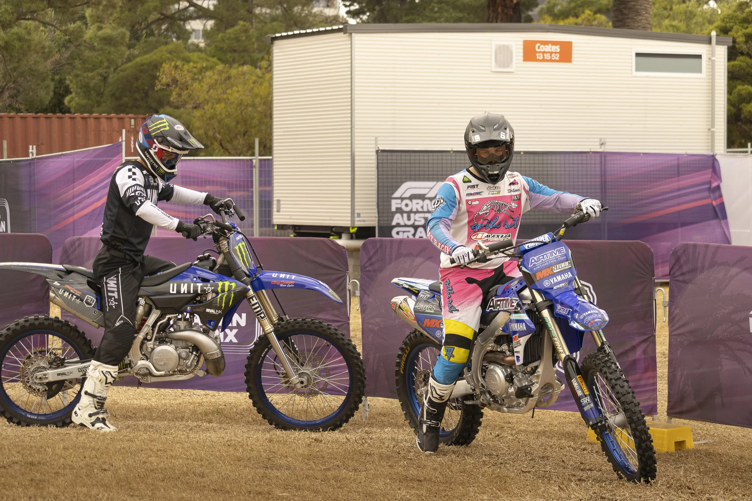 Two motocross riders in full gear and helmets on dirt bikes, preparing on a race track with purple barriers and a container in the background.