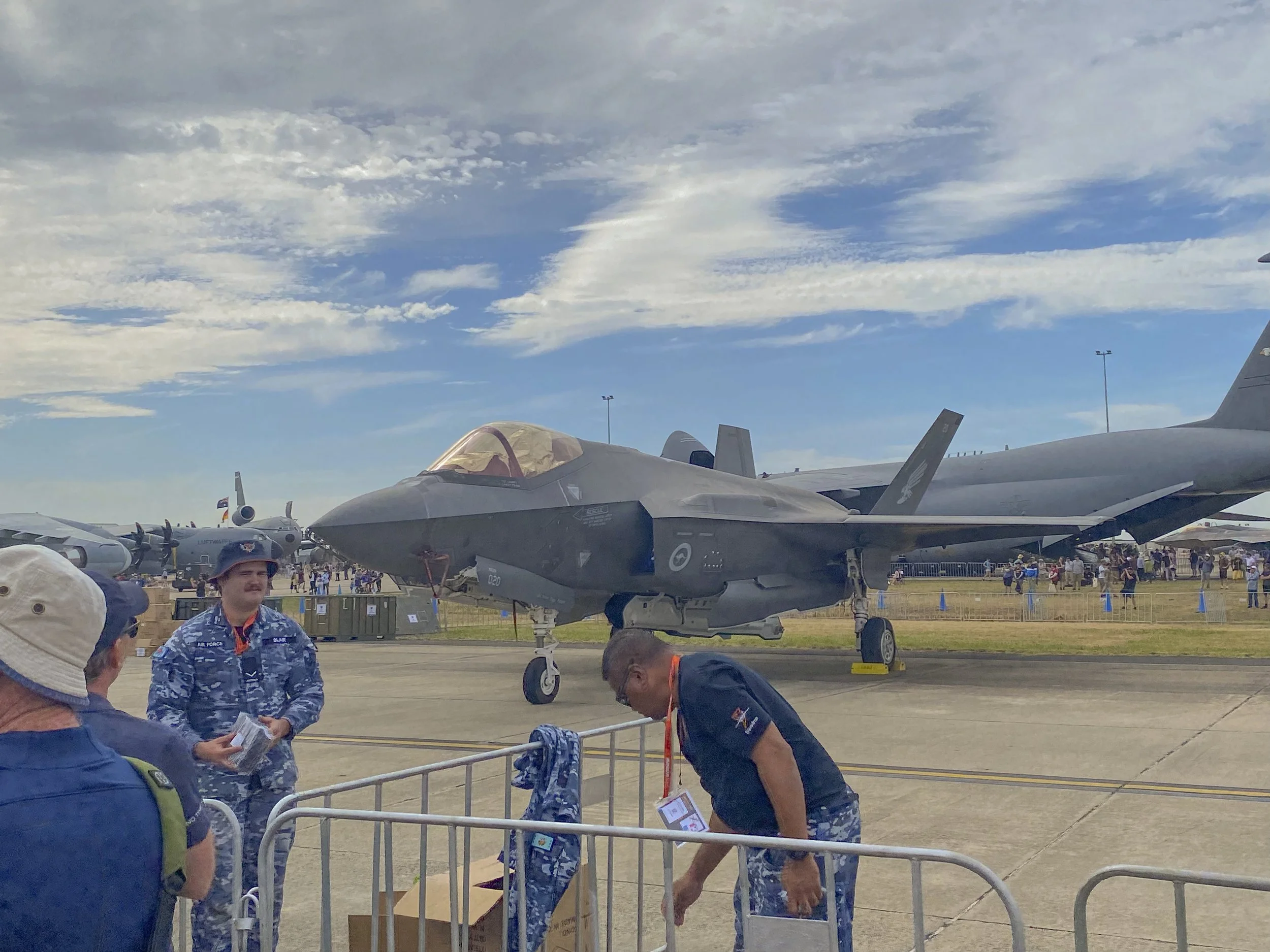 A military airshow with a fighter jet on display. There are several people, including uniforms and civil attire, standing around the jet. The background shows other aircraft and a crowd, under a partly cloudy sky.