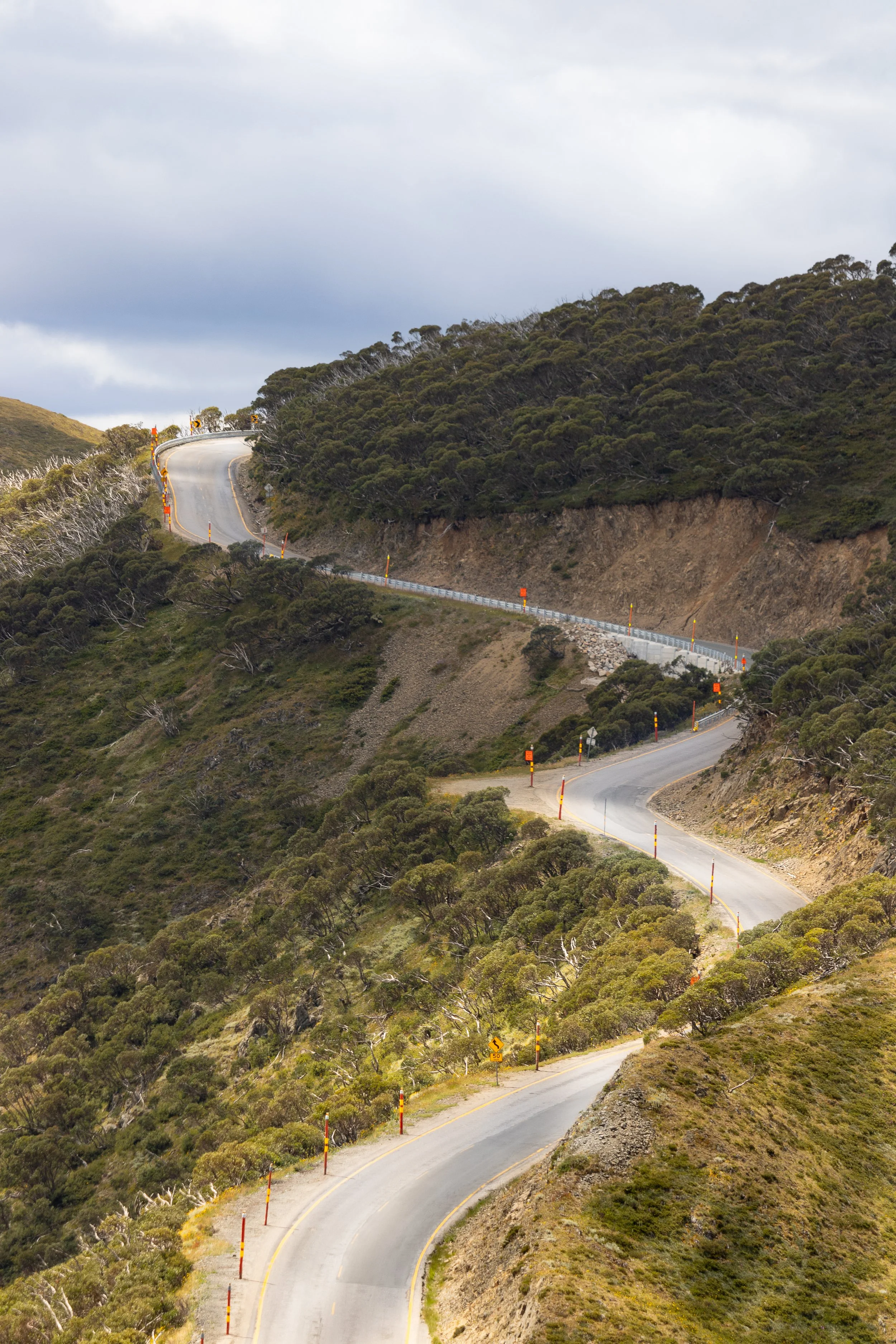 Winding mountain road with guardrails, yellow and orange markers, surrounded by green shrubbery and hills under a cloudy sky.