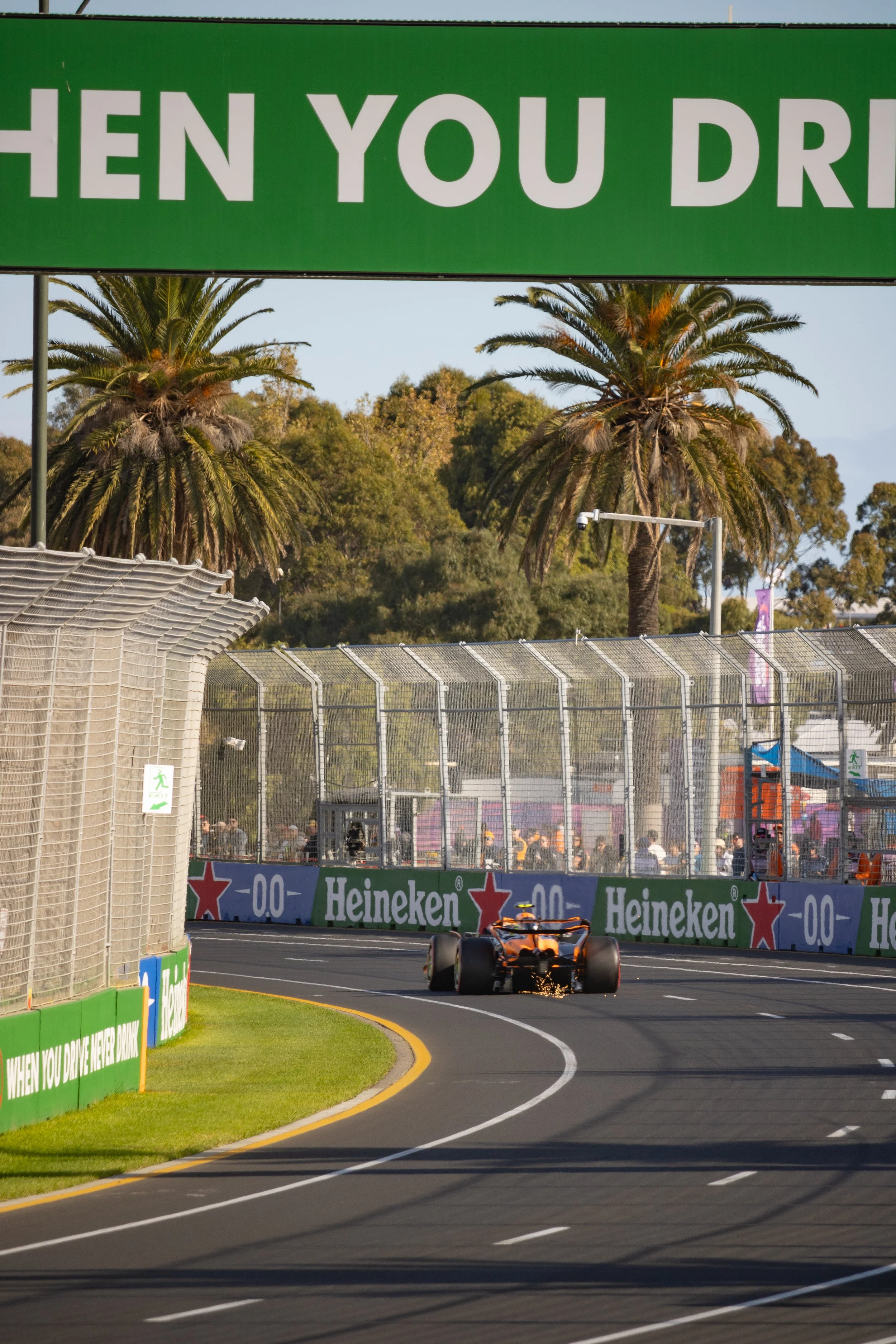 A race car drives on a road near a barrier with Heineken advertisements during a racing event, with palm trees and spectators in the background.