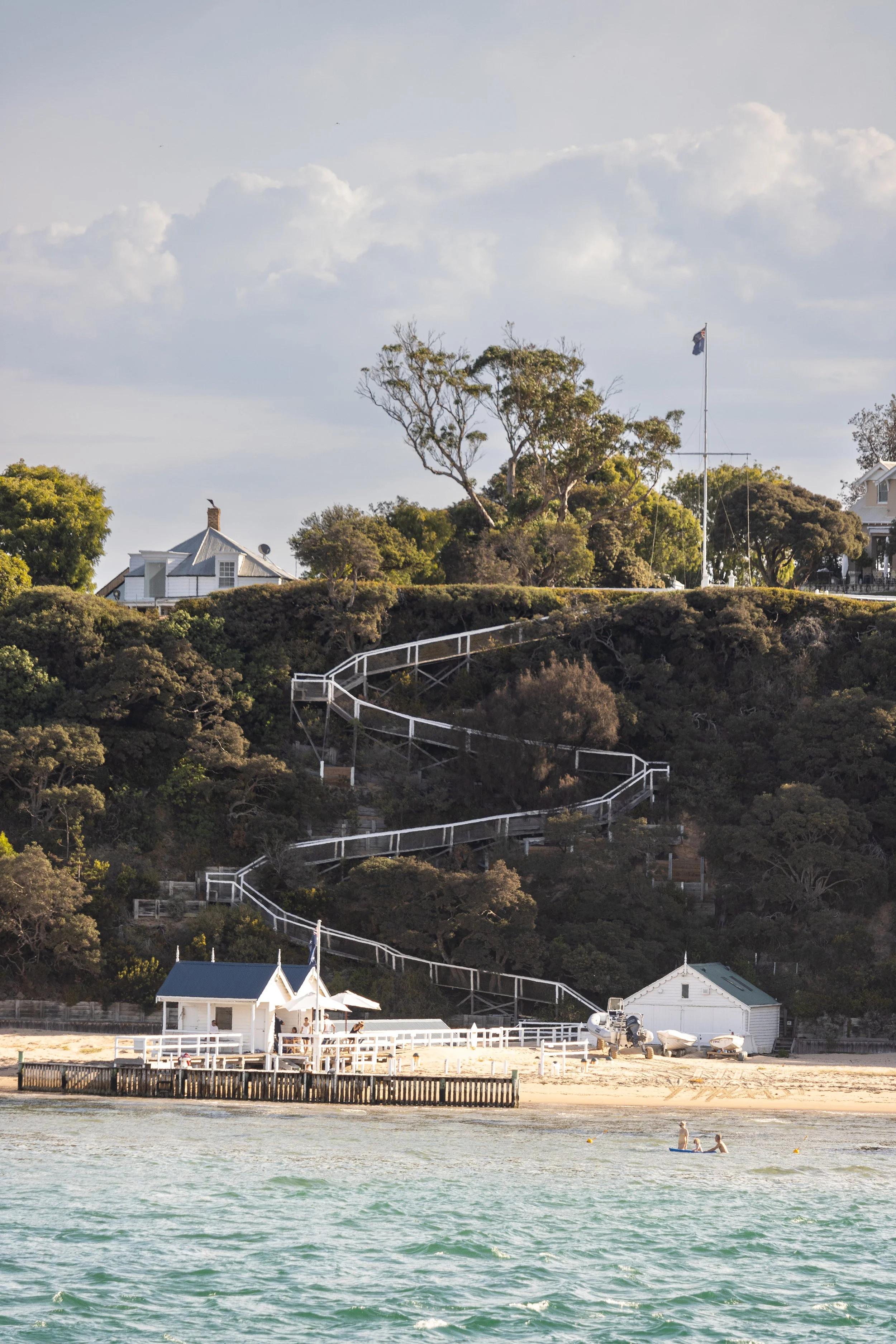 Scenic view of a beach with a small white building, a pier, and people swimming; a hillside with trees and white buildings with a spiral staircase and flagpole in the background.