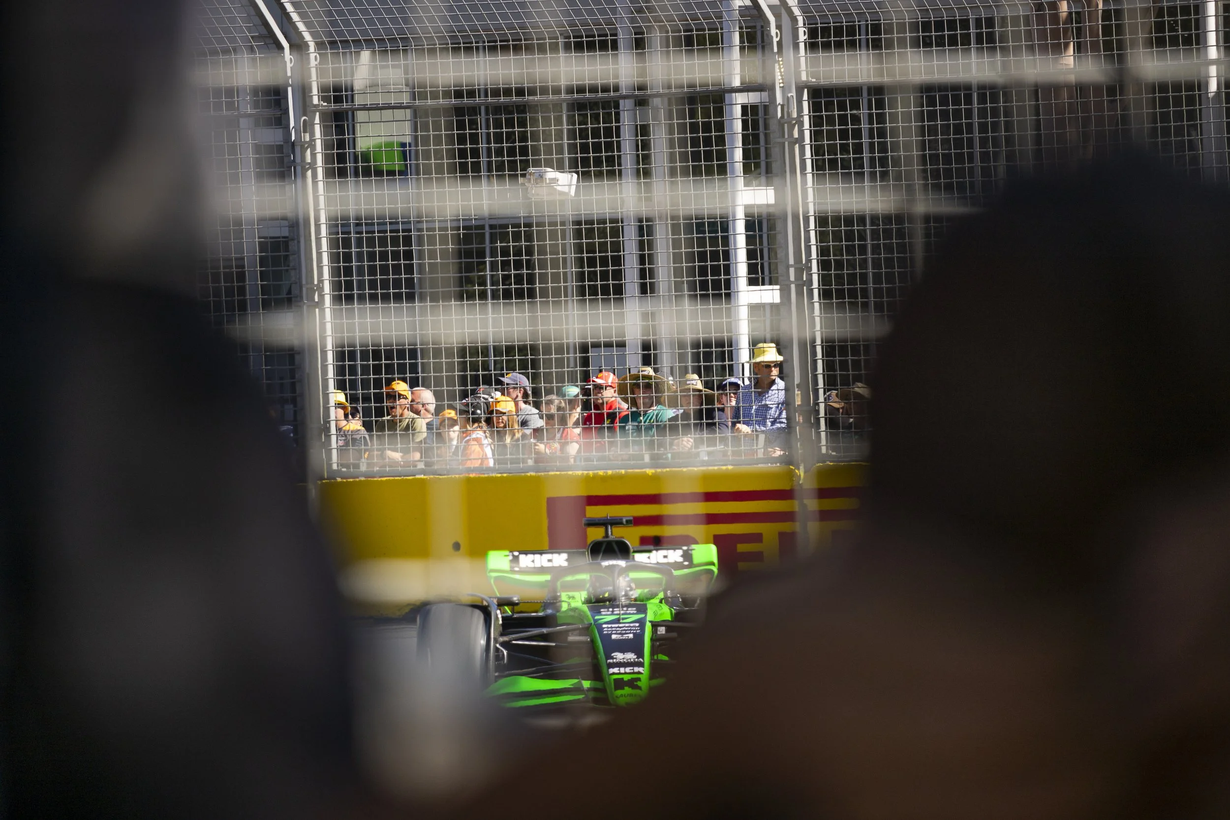 A green and black racing car on the track viewed through safety barriers, with spectators watching behind the barriers.