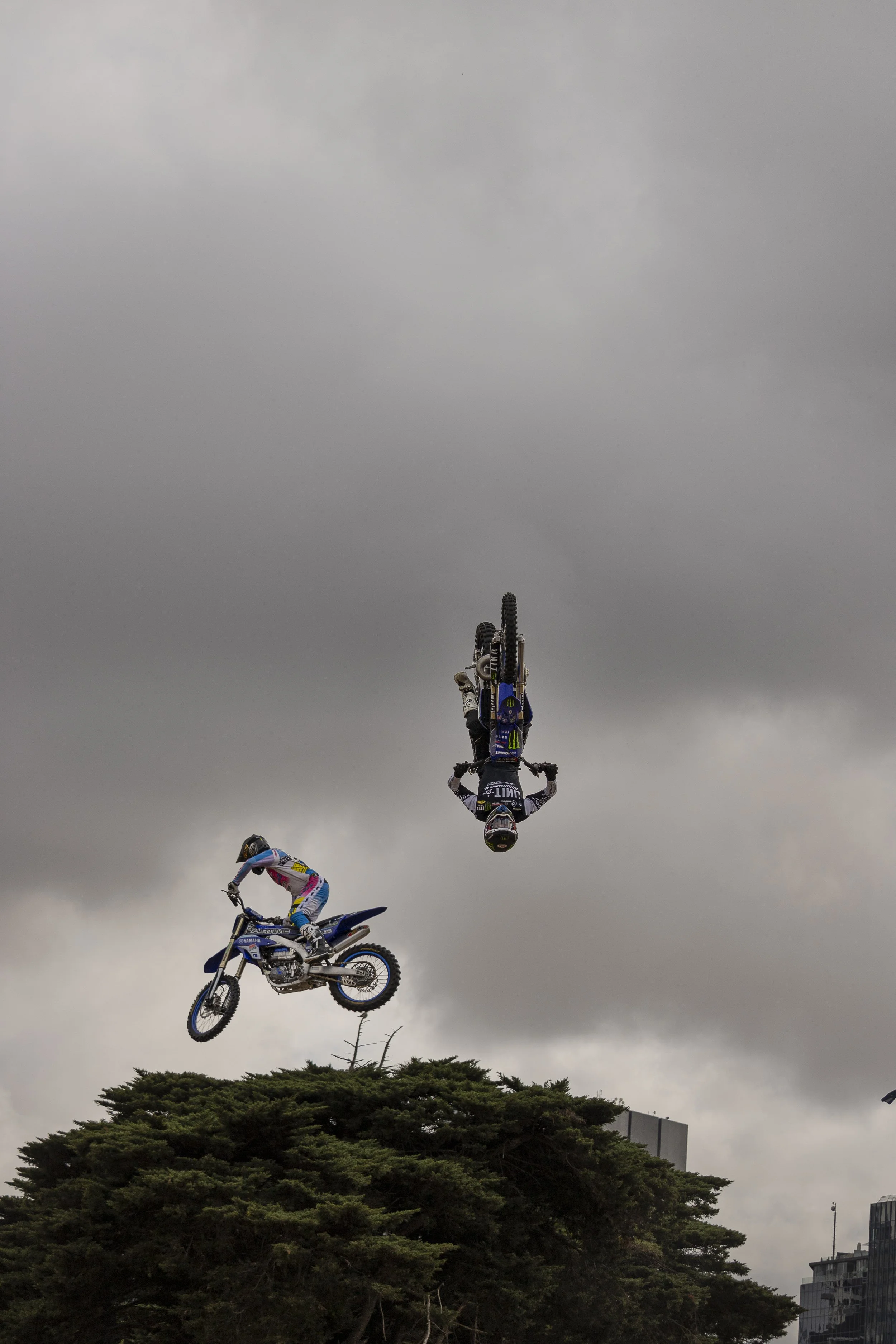 Two motocross riders performing aerial stunts over trees against a cloudy sky, with one rider upside down and the other mid-air.