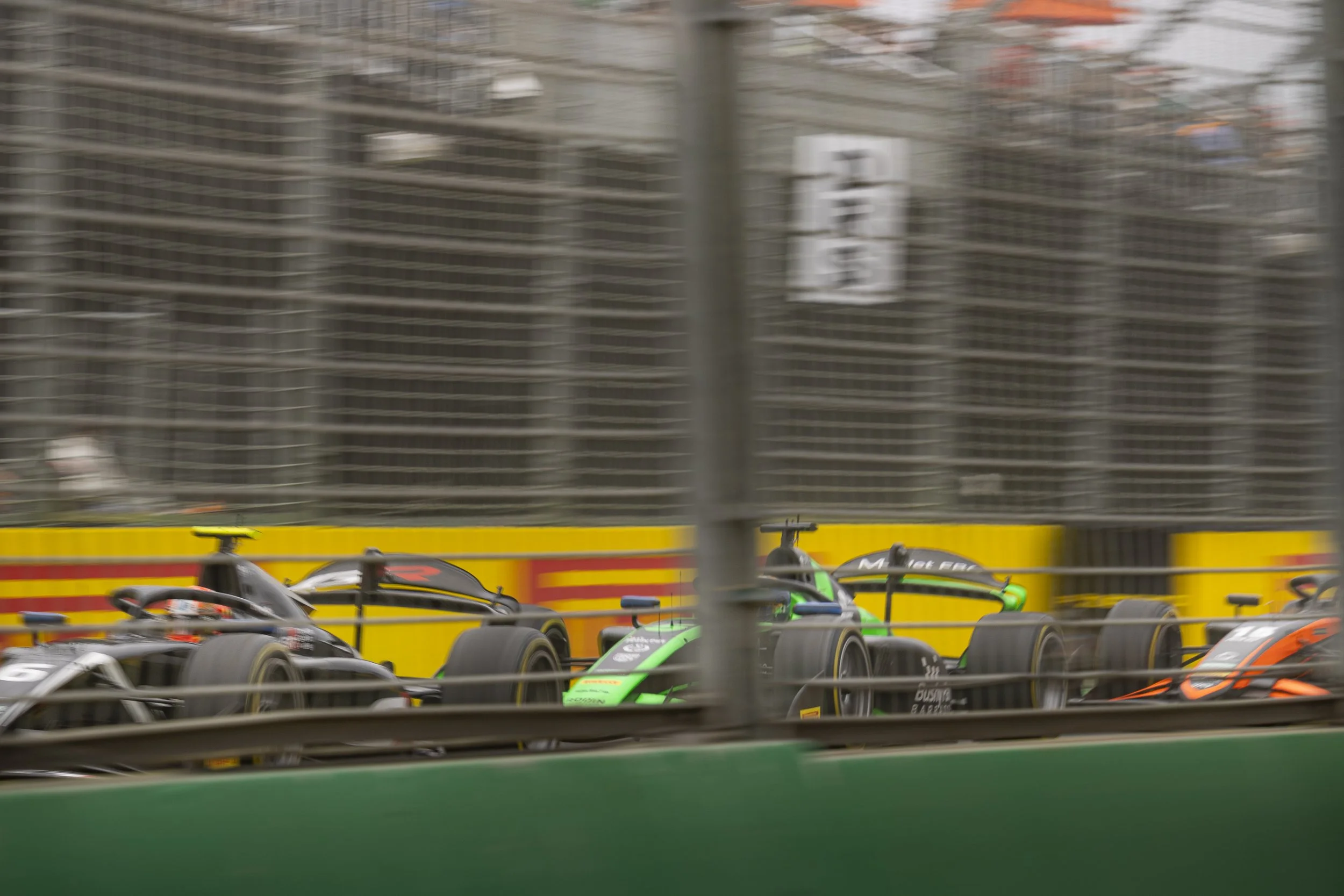 Race cars on a track with barriers and fencing, blurry background showing city buildings.