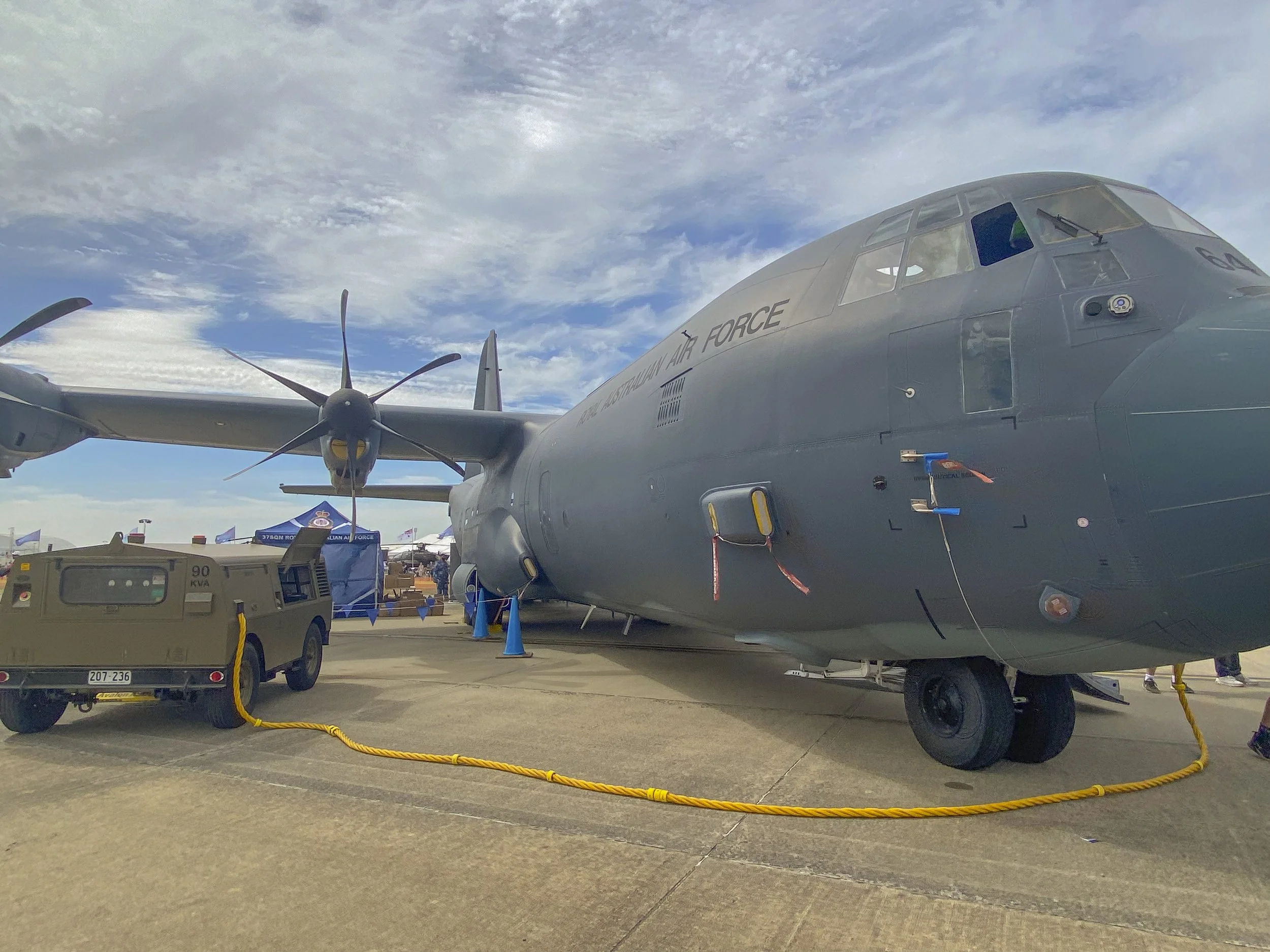 A large military aircraft on display at an air show, with a vehicle connected to its side and a blue sky with clouds overhead.