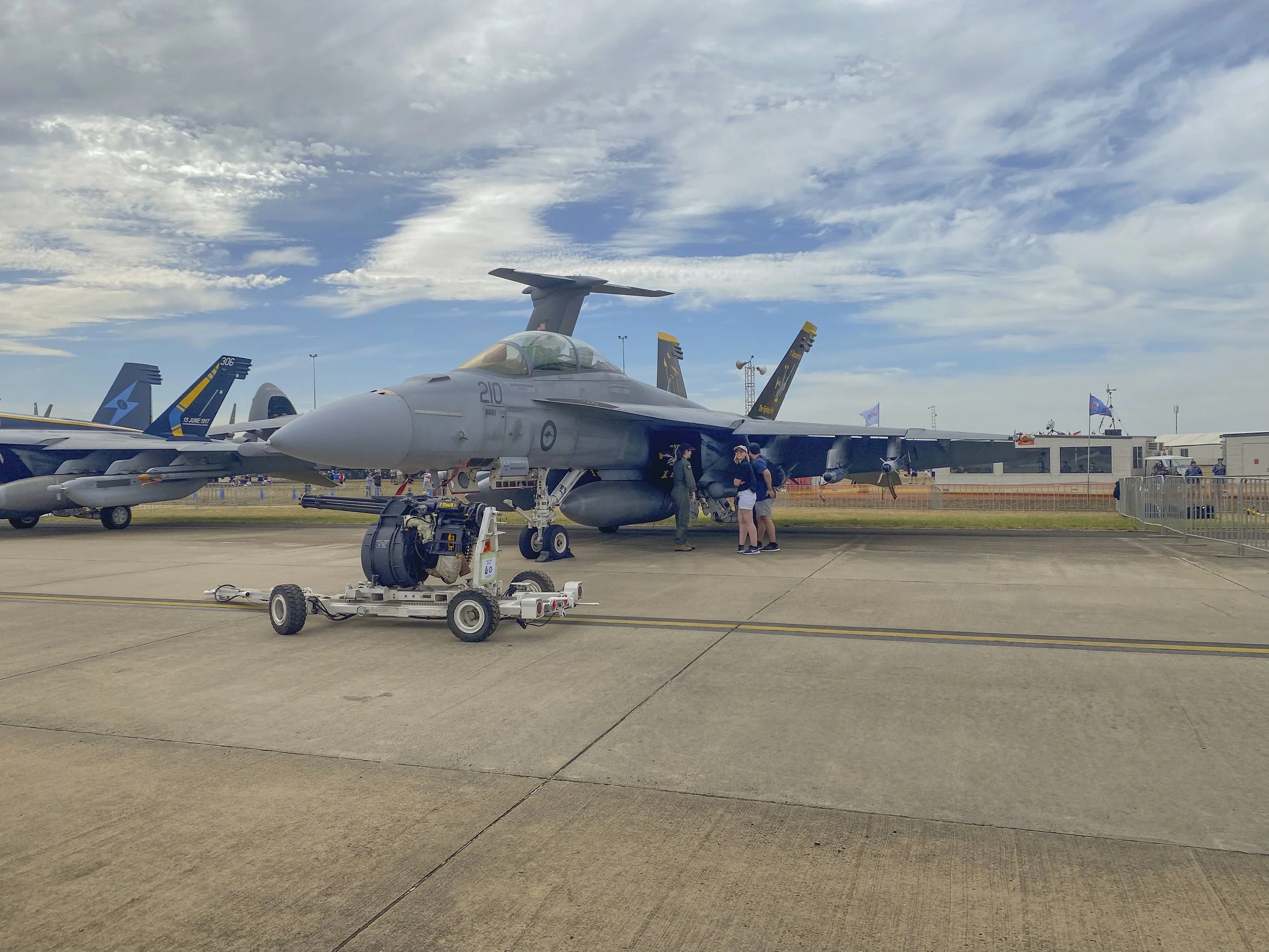 A fighter jet aircraft on display at an air show, with people gathered around it, and other aircraft visible in the background under a partly cloudy sky.