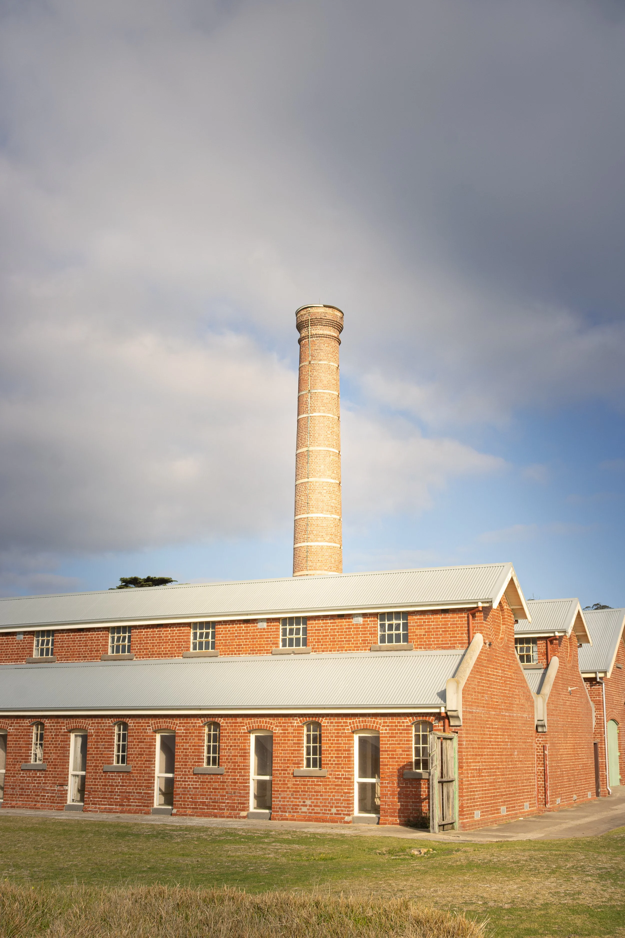 A brick building with multiple small windows and a tall smokestack chimney under a cloudy sky.