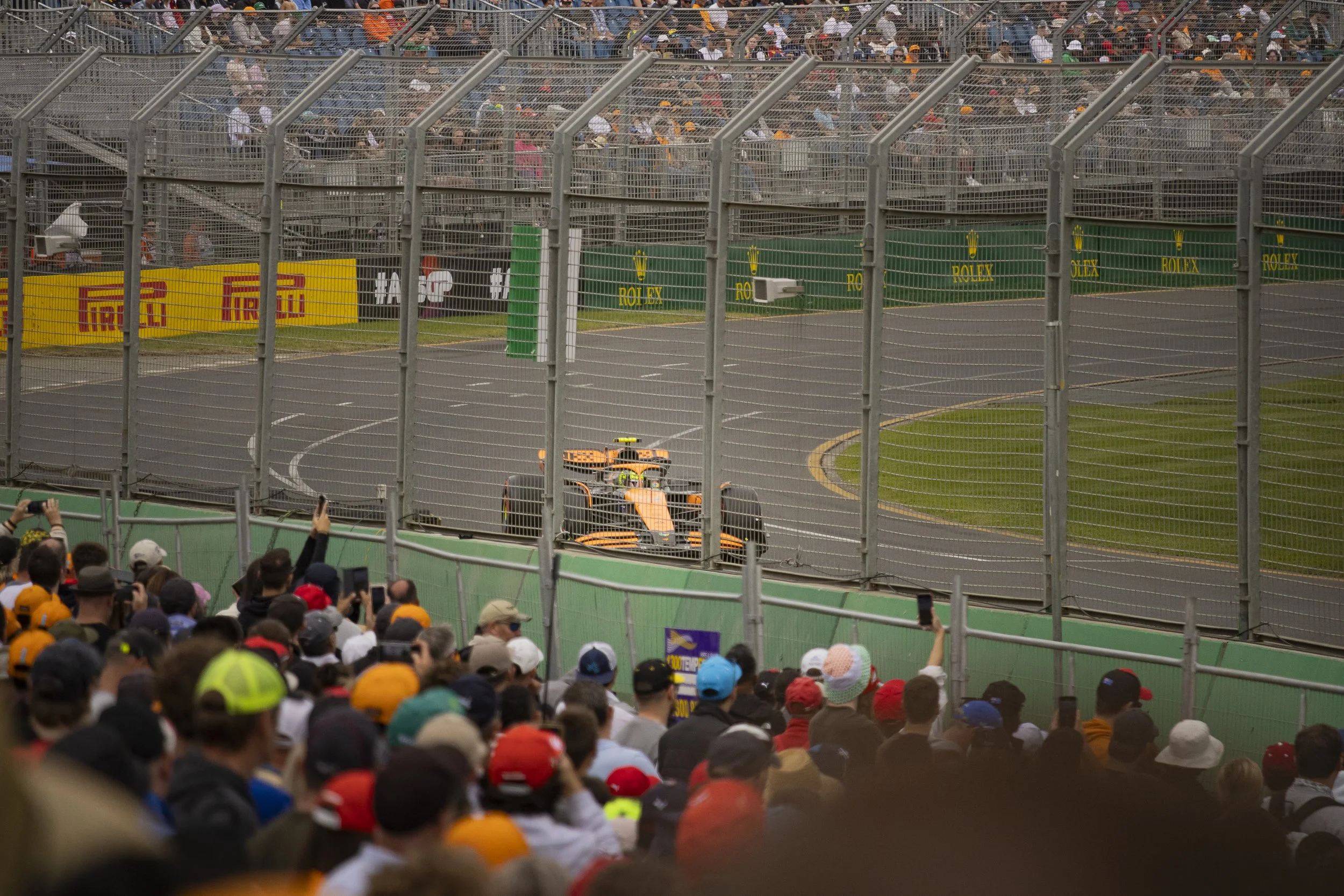 Race car driving on track during a race, spectators watching from the stands and taking photos, surrounded by safety barriers and advertisements.