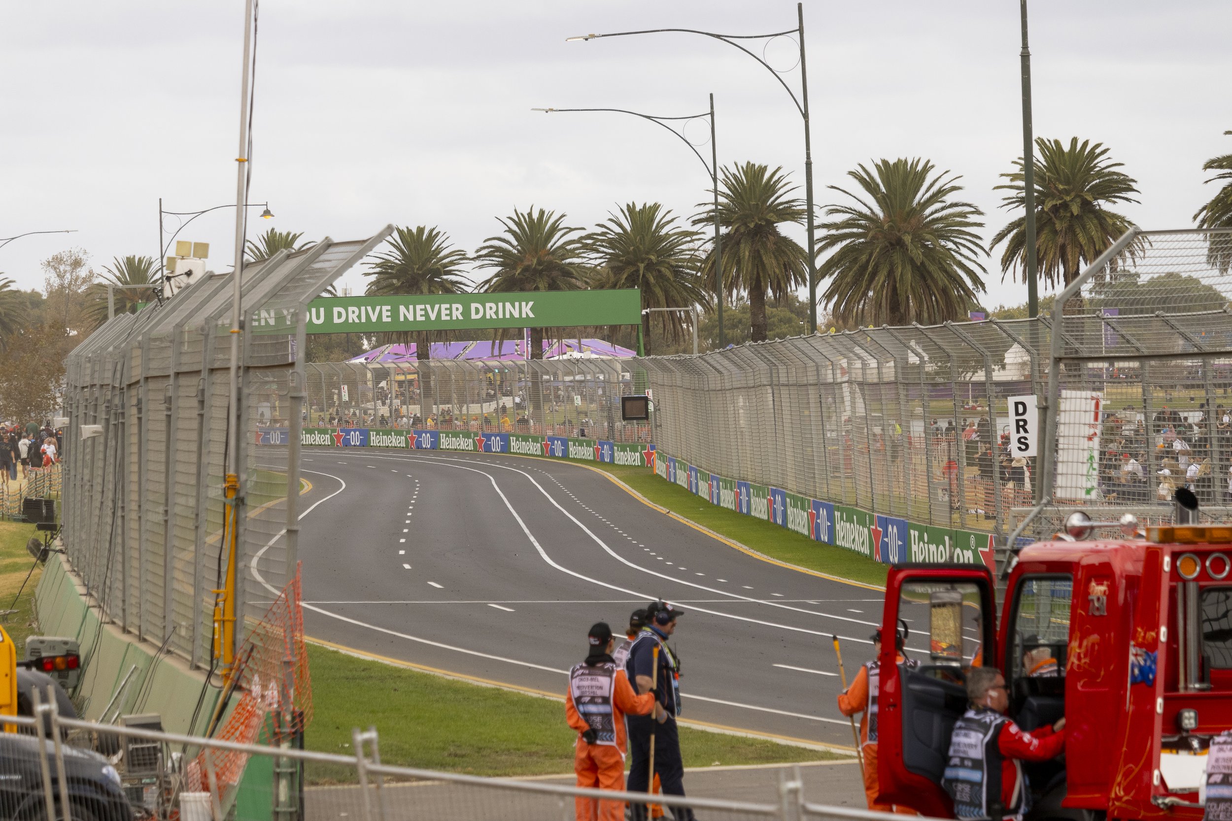 View of a race track with safety fences, palm trees in the background, and caution personnel near a red fire truck.