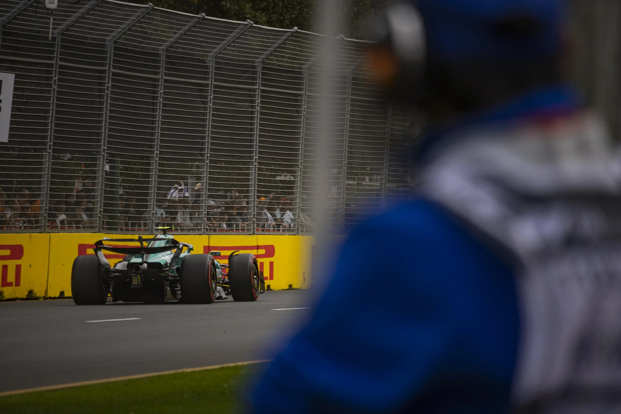 A race car on the track with a person in racing gear in the foreground, spectators behind the safety barrier, and a yellow advertisement board.