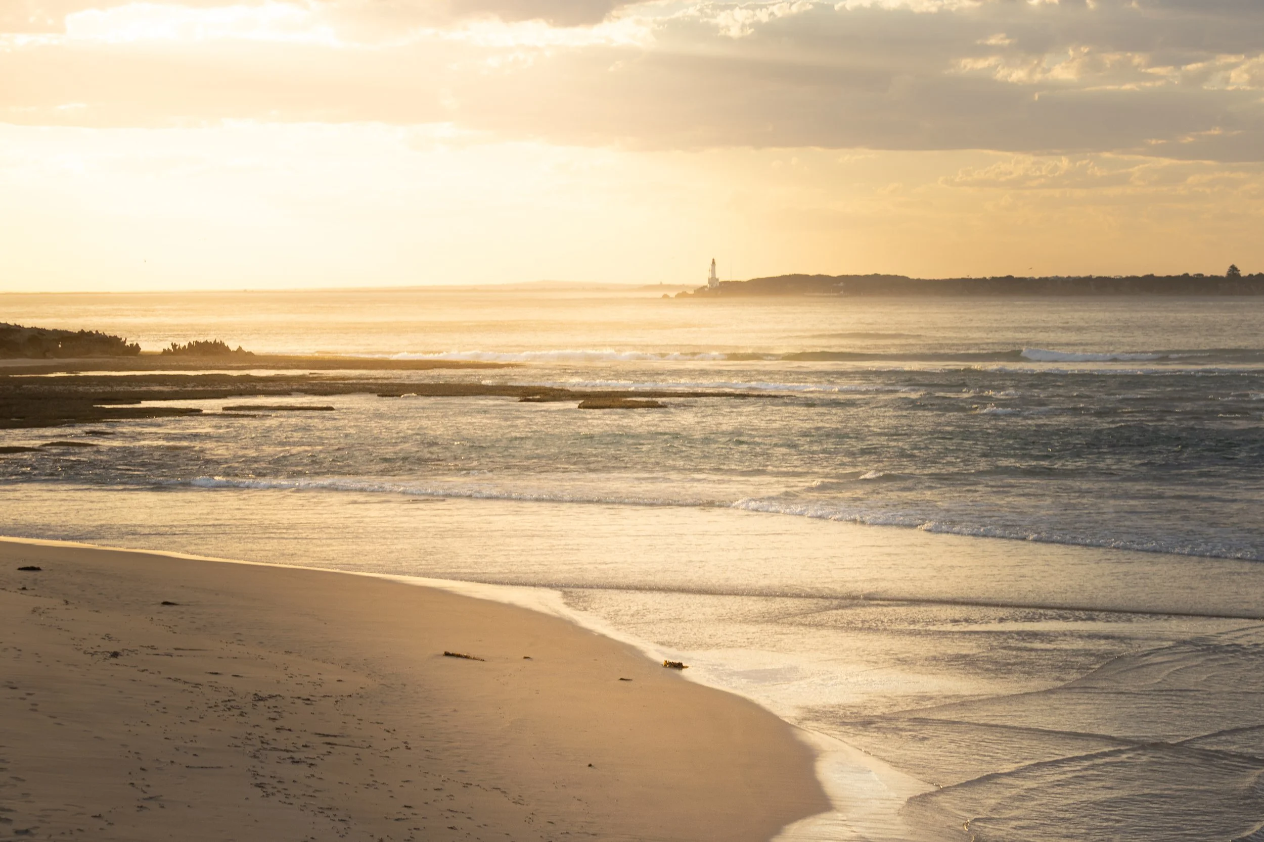 A serene beach scene during sunset with calm waves, sandy shore, and a distant land with a lighthouse.