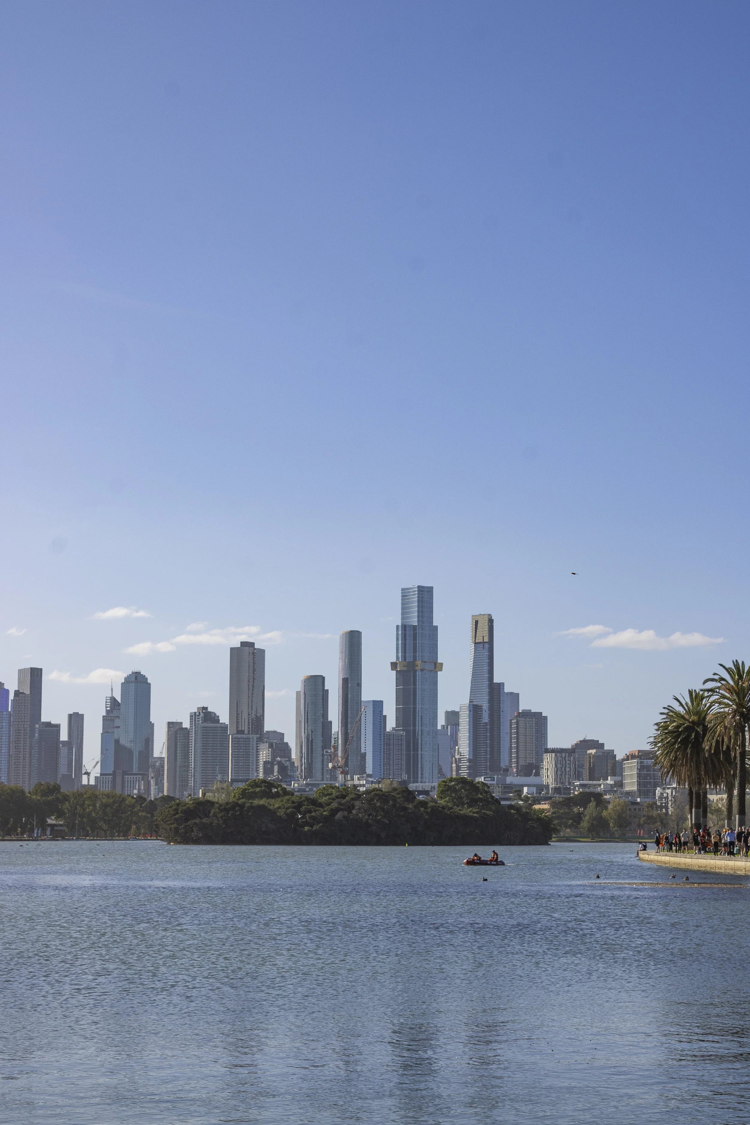 A city skyline with tall skyscrapers seen from across a body of water, with a small boat and some people on the right side near the water.