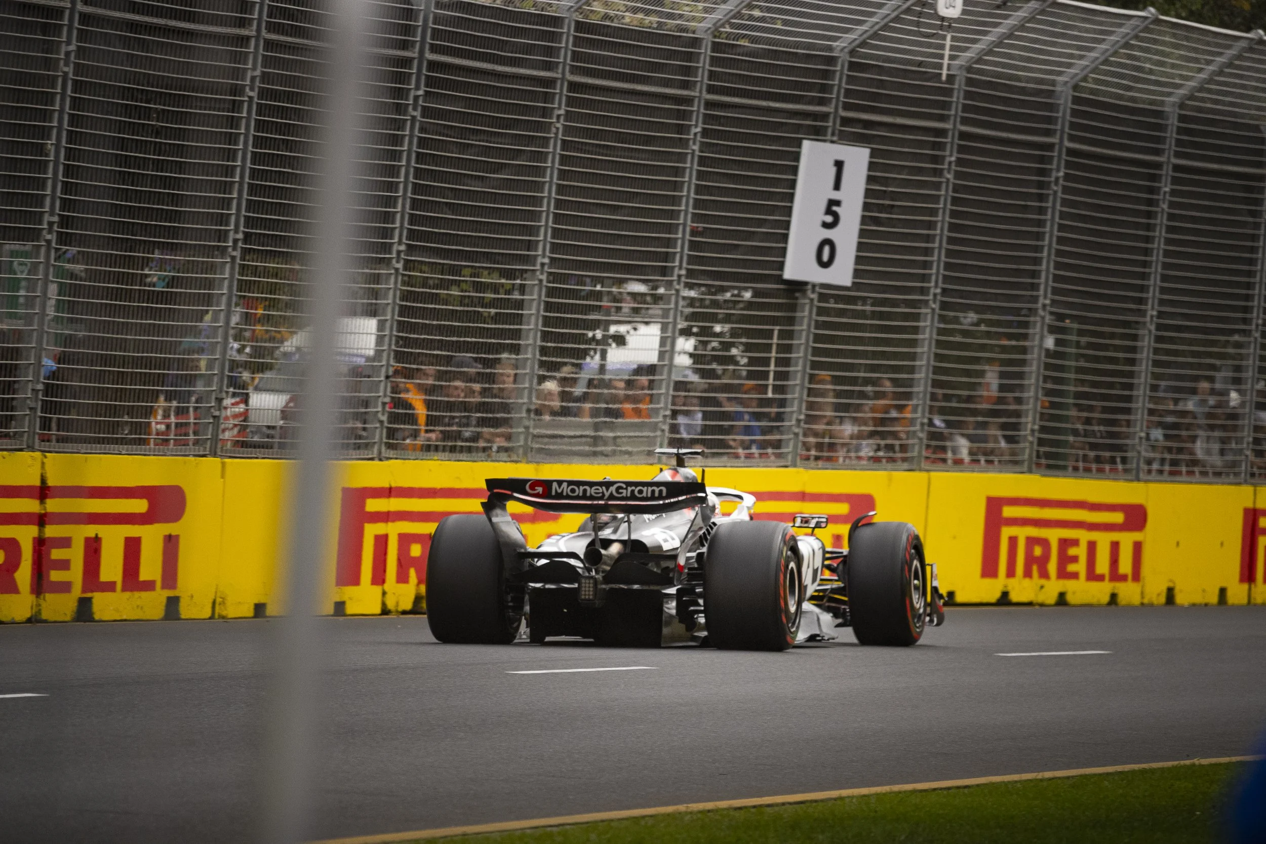 Formula 1 race car on track with Pirelli and MoneyGram logos, viewed through a metal fence, with spectators behind.