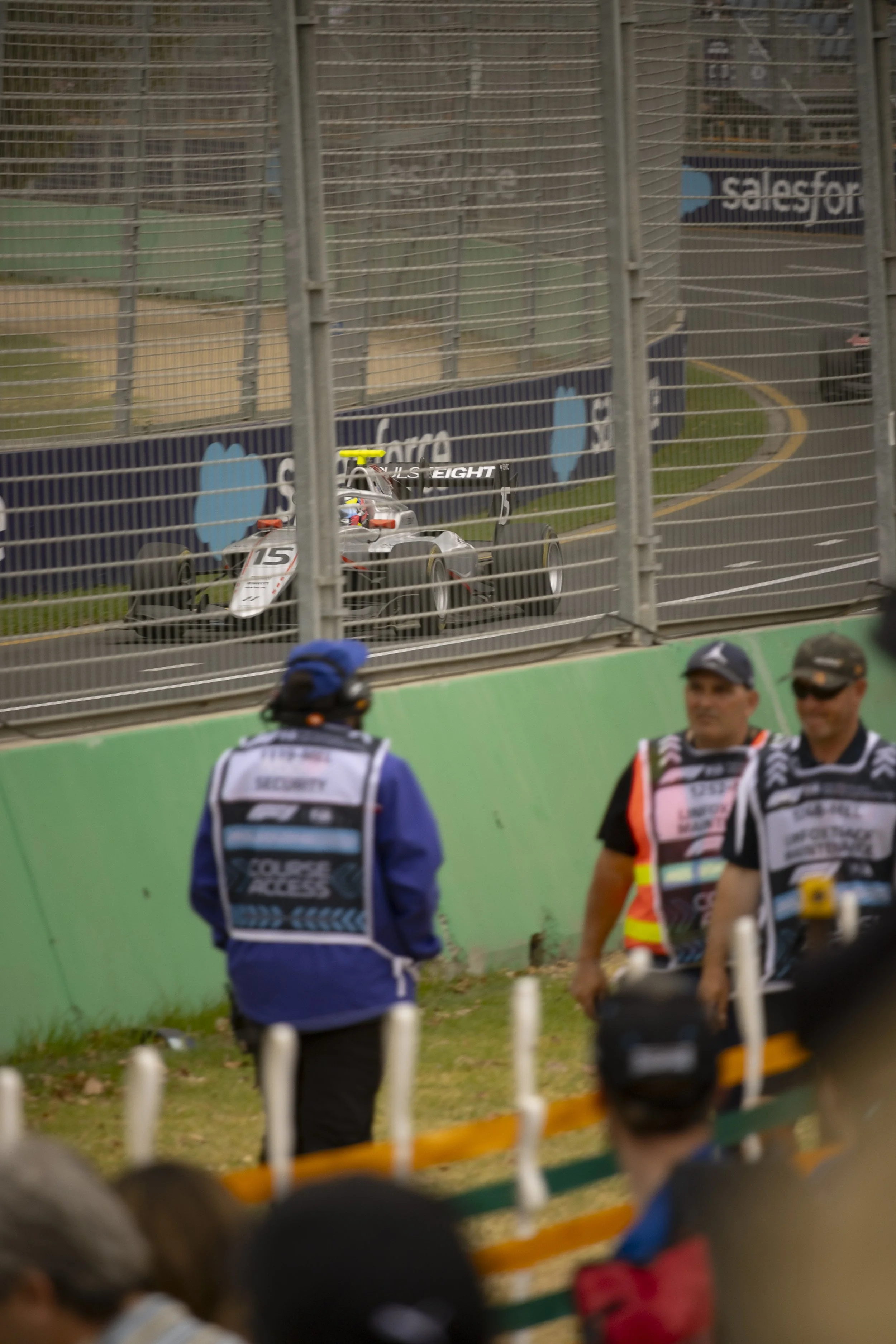 A race car on a track seen through a safety fence, with people watching and officials standing nearby.