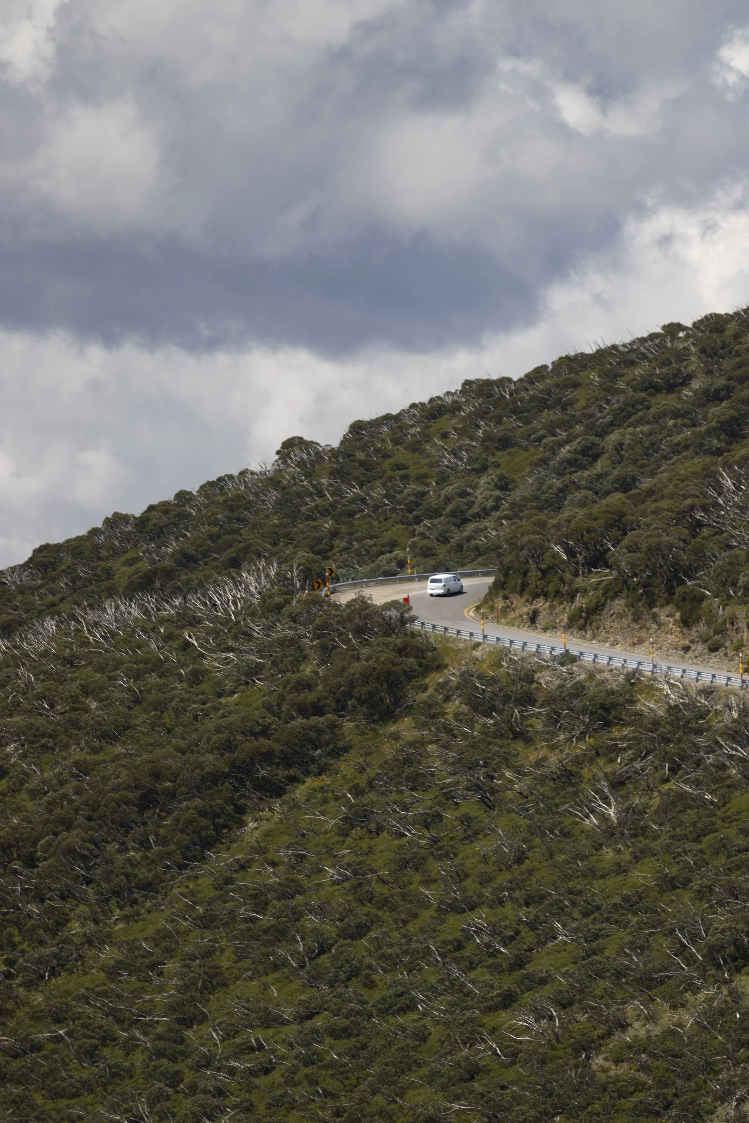 A white car driving on a winding mountain road surrounded by green trees, under a cloudy sky.