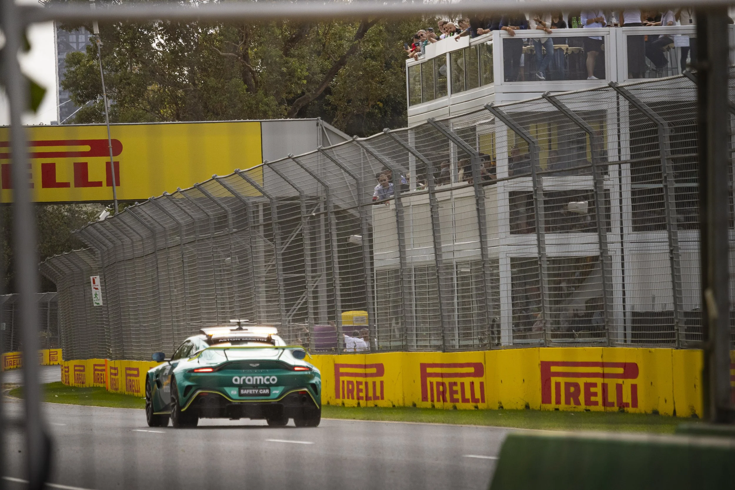 Race car on a wet track with Pirelli advertising barriers. Spectators watching from a viewing platform above the track.