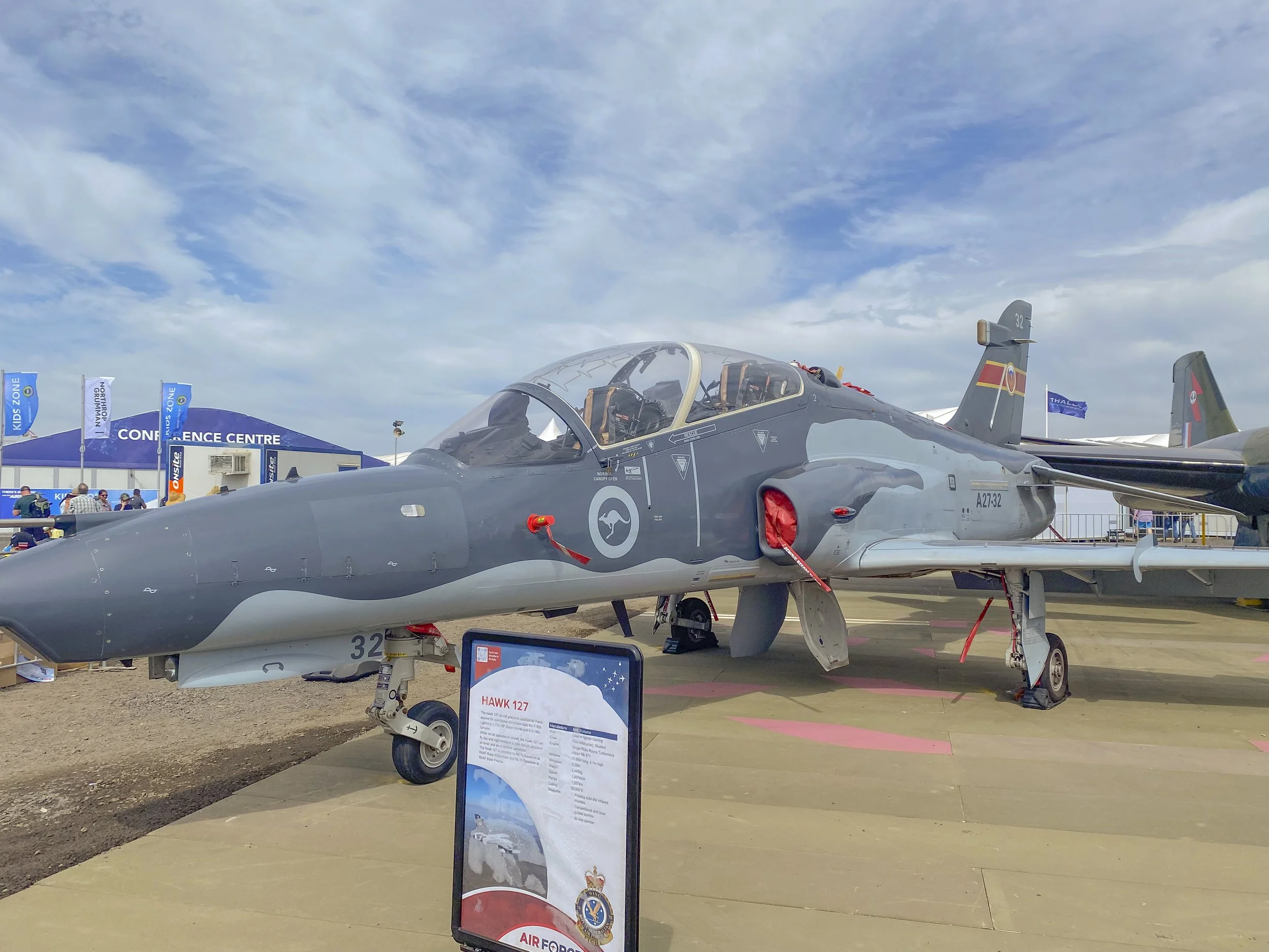 A military fighter jet on display at an airshow with a sign providing information in front of it, and a conference center building in the background with blue flags, under a partly cloudy sky.