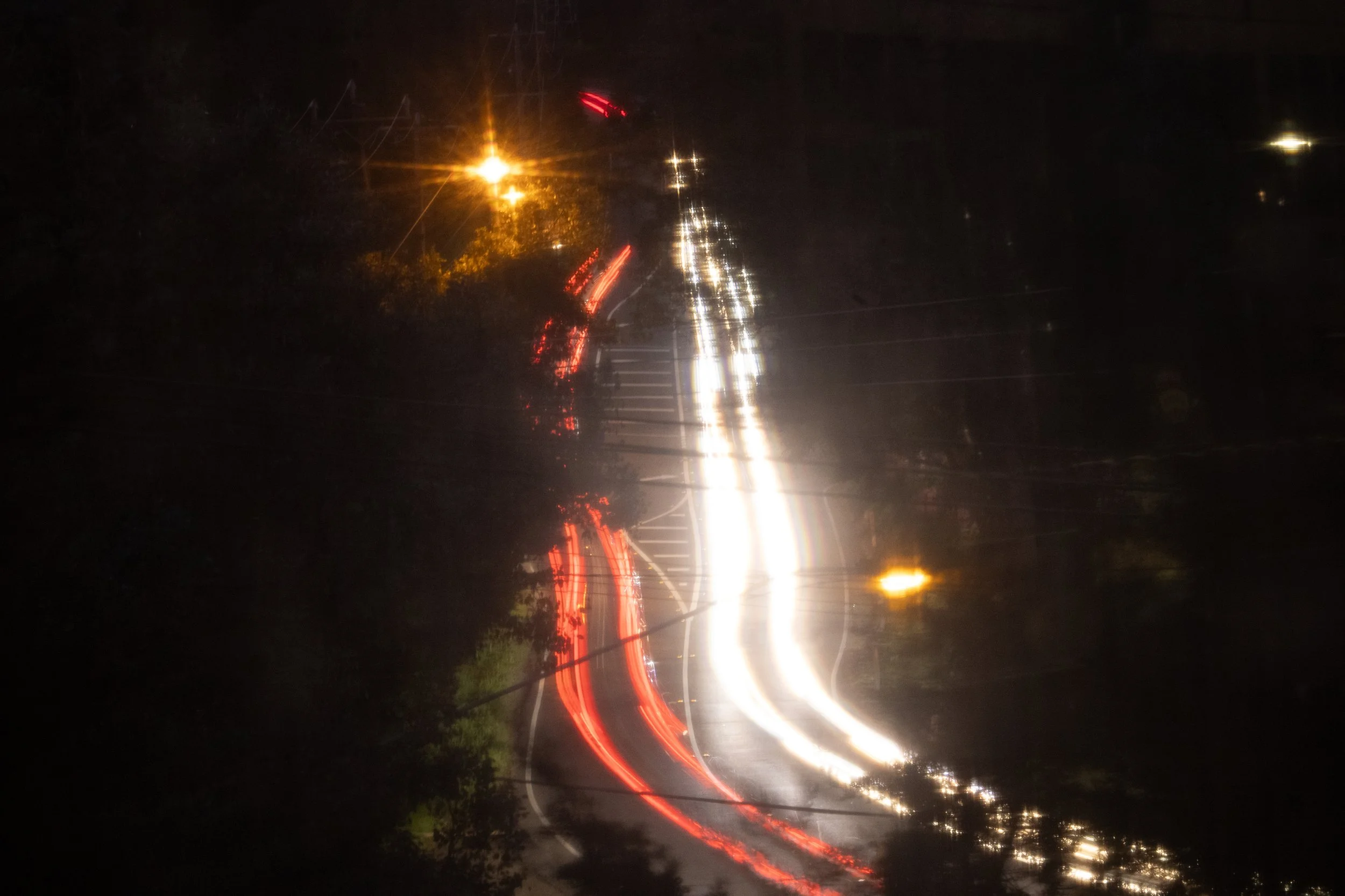 Nighttime scene of a wet road with car headlights and taillights creating streaks of light due to long exposure, with trees and streetlights visible in the background.