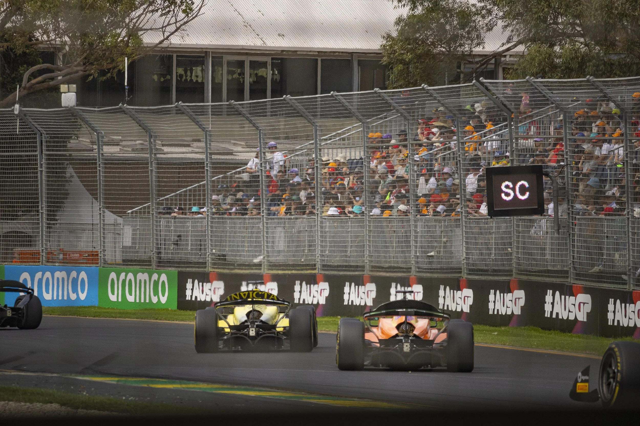 Race cars on a track with spectators behind a safety fence, and digital display showing 'SC' for safety car.