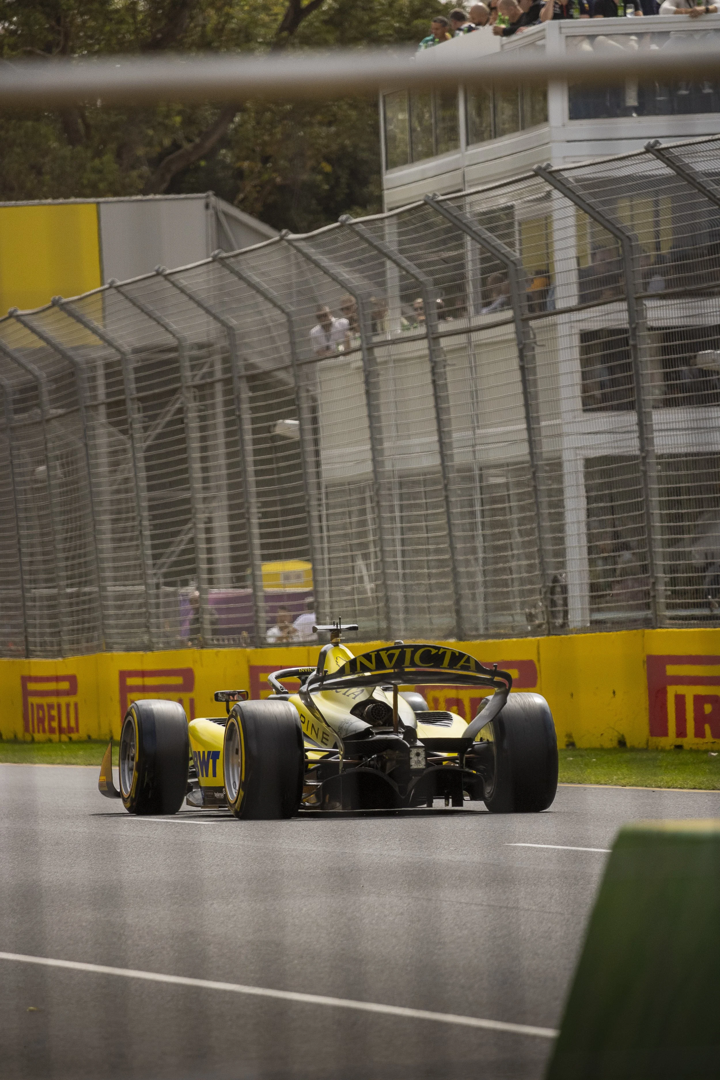 A yellow and black IndyCar racing car on a race track near the audience stand and safety barriers, with people watching from the upper level.