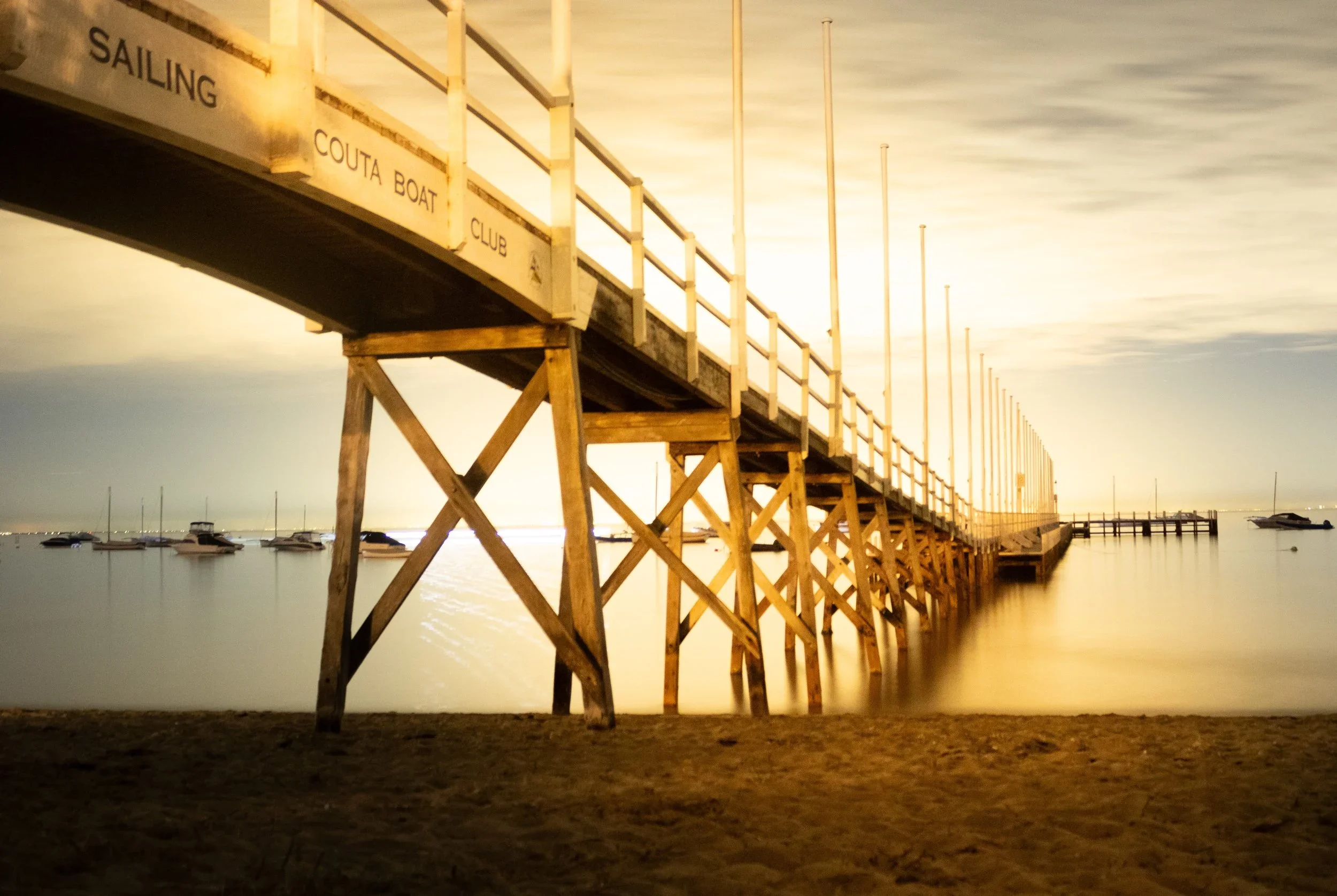 A wooden dock extending into a calm water body with boats anchored nearby, under a cloudy sky with warm lighting.