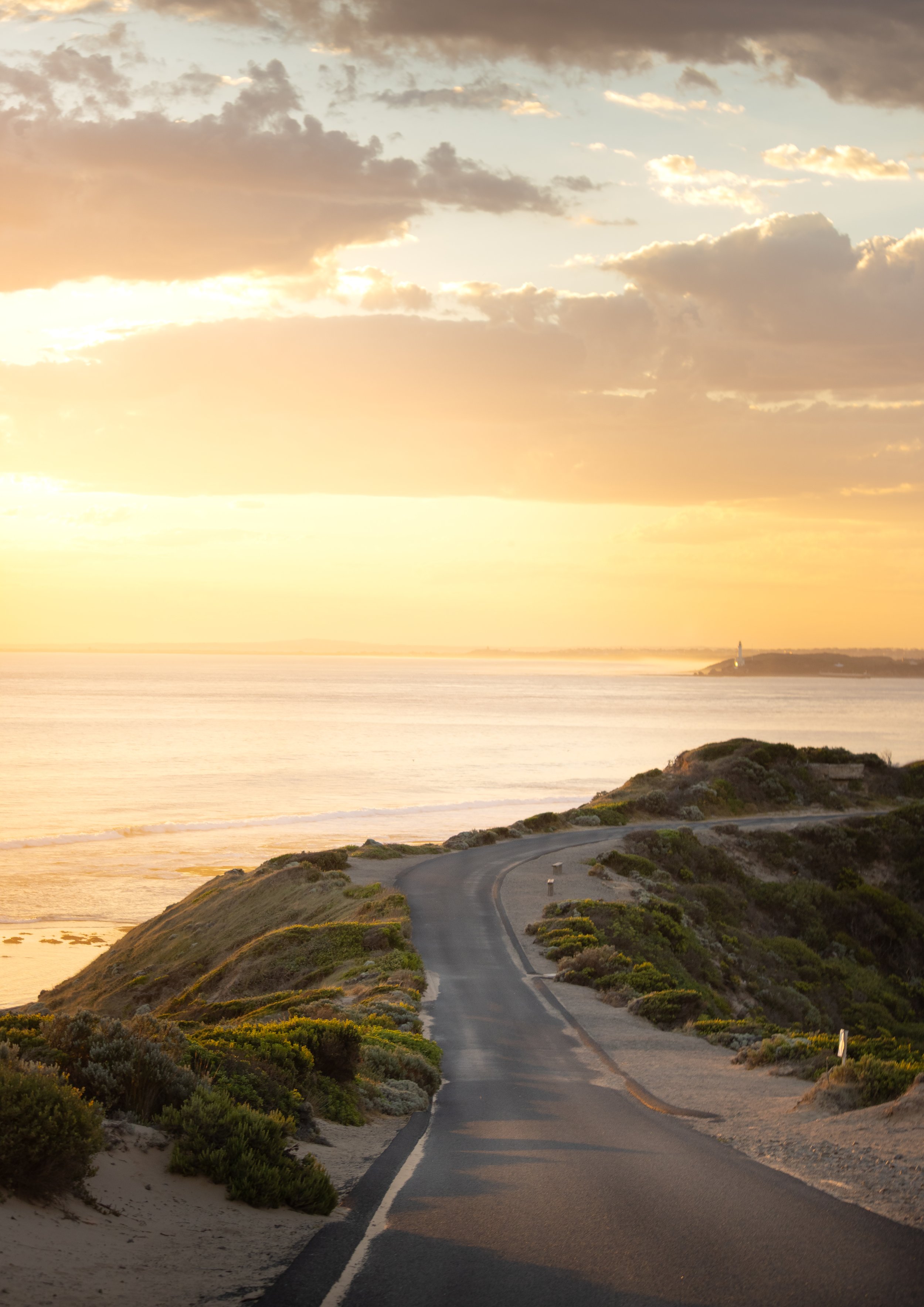 A winding coastal road at sunset with a view of ocean and a lighthouse on a distant headland. The sky has clouds with golden sunlight.
