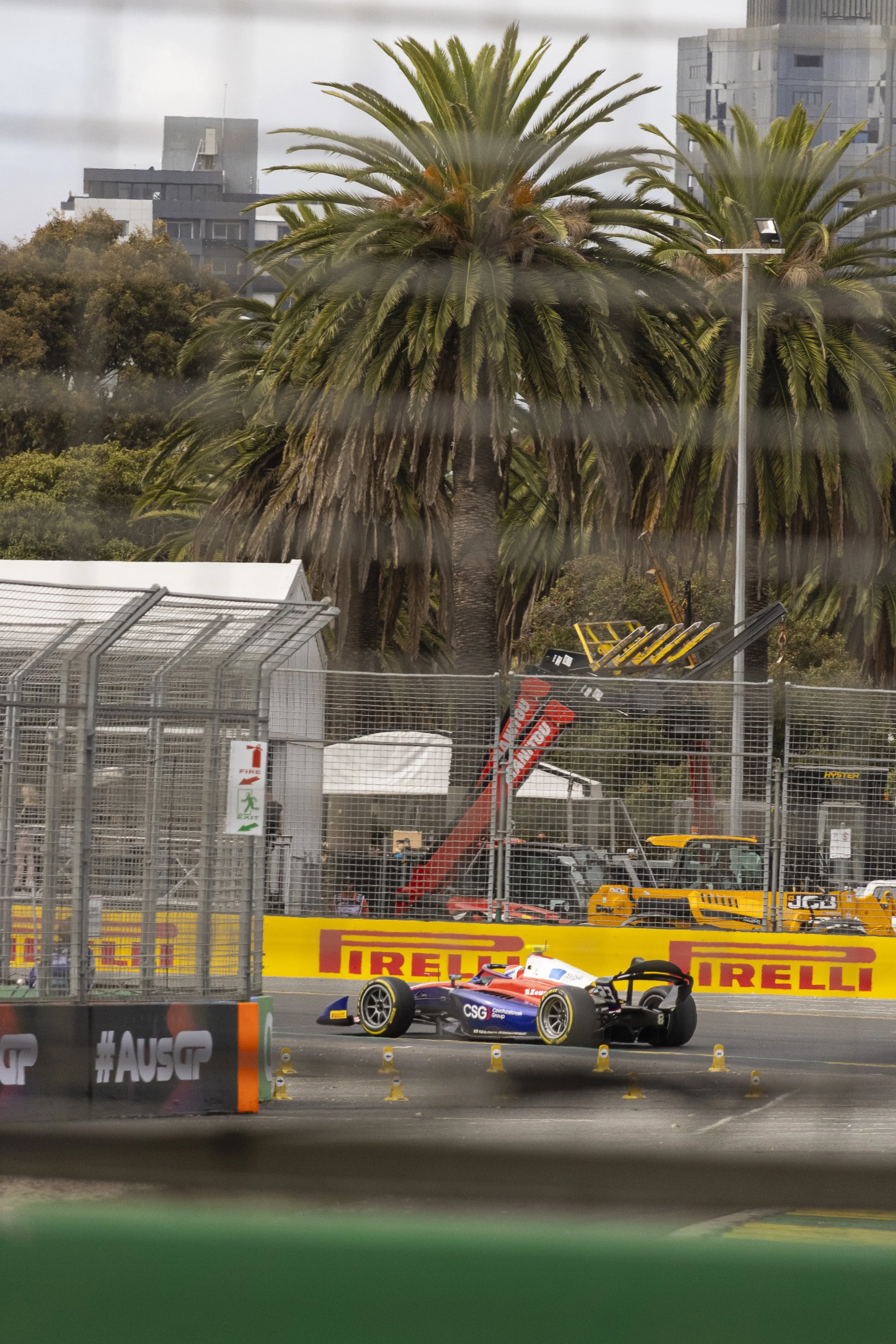 Race car on the track with palm trees and construction equipment in the background, seen through a rain-covered window.
