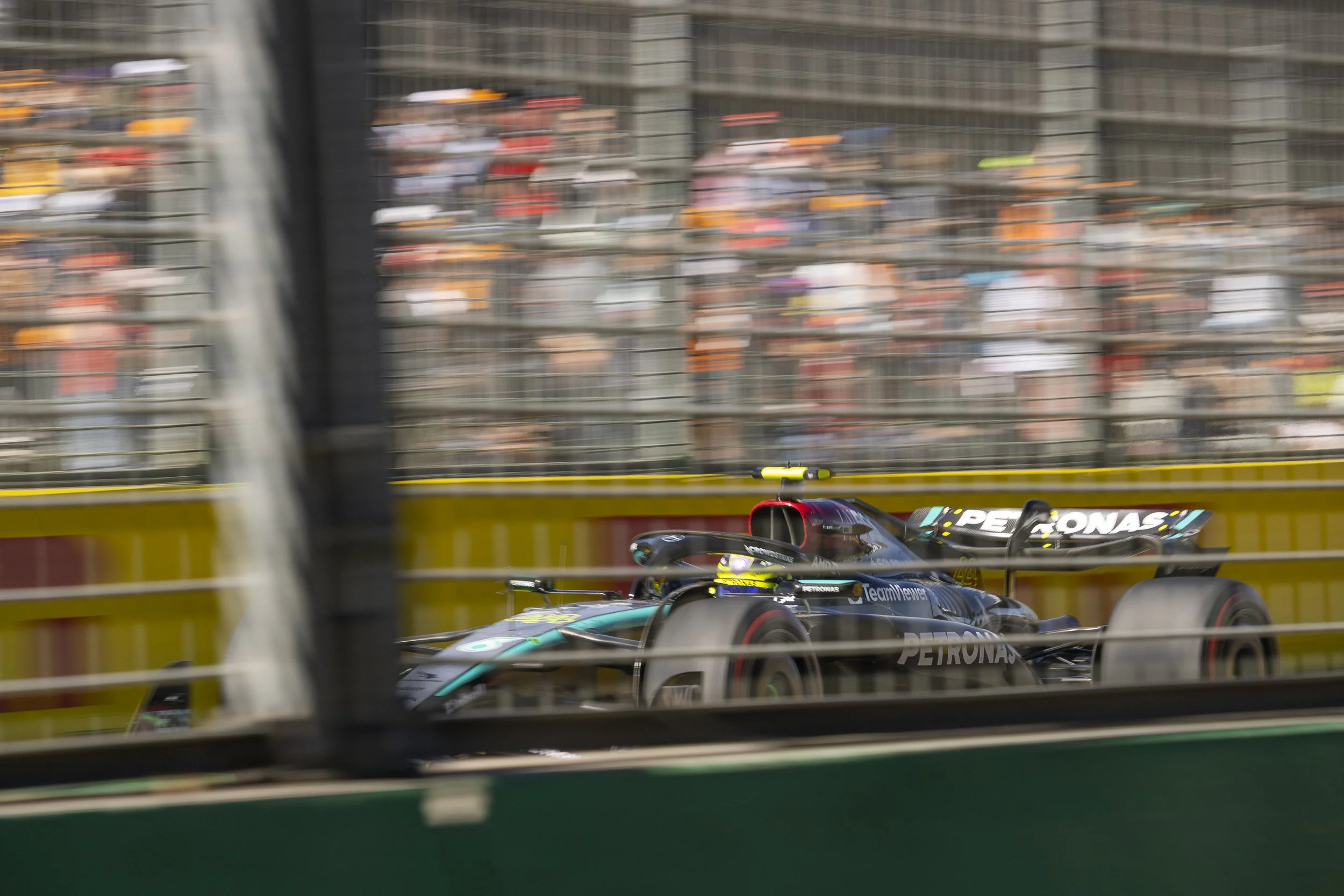 A racing car on a track visible through a fence, with a blurred background of spectators in the stands.
