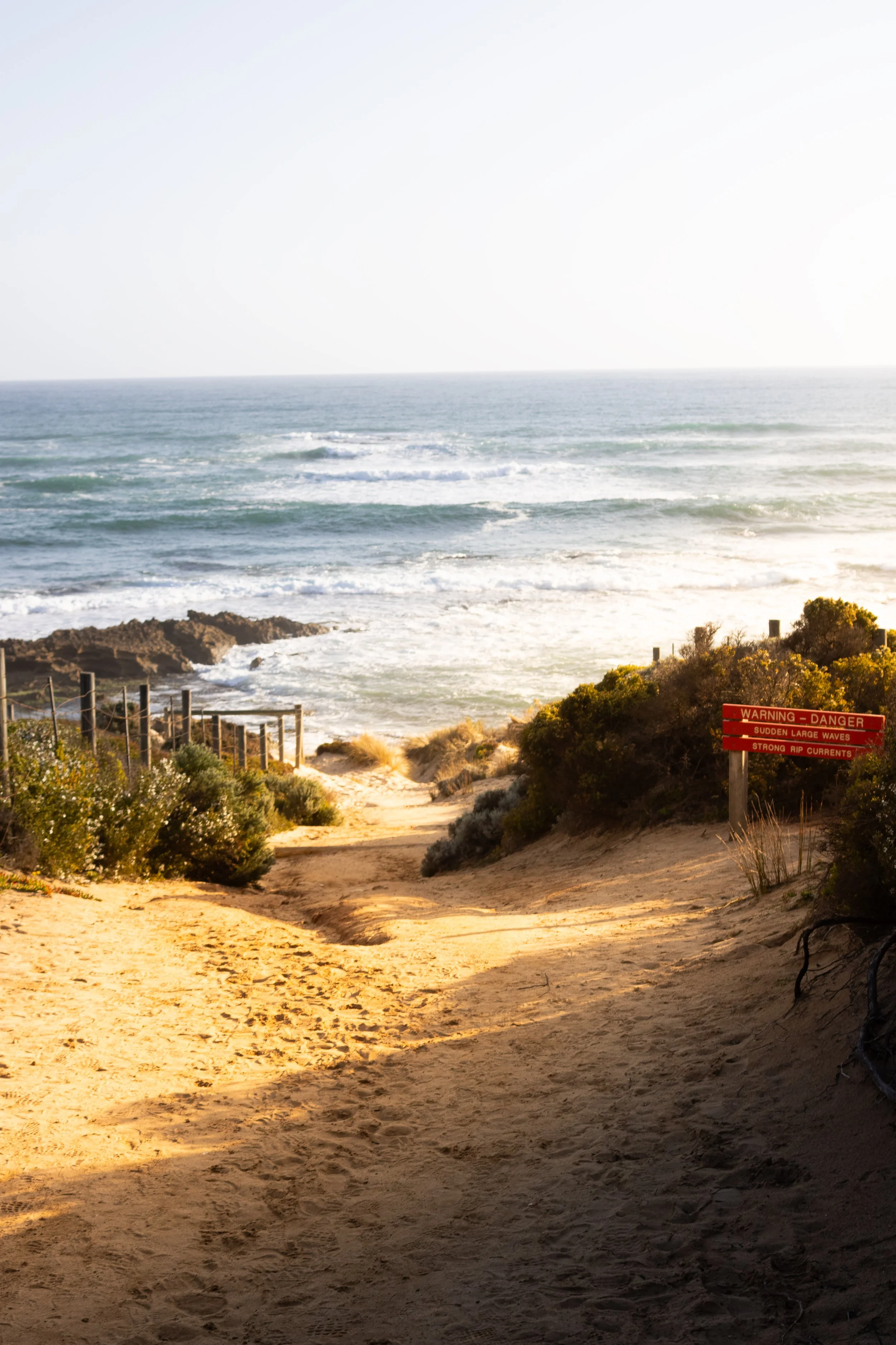 Dirt pathway leading down to the beach with warning signs about large waves and strong currents, flanked by bushes and fencing, with the ocean in the background.