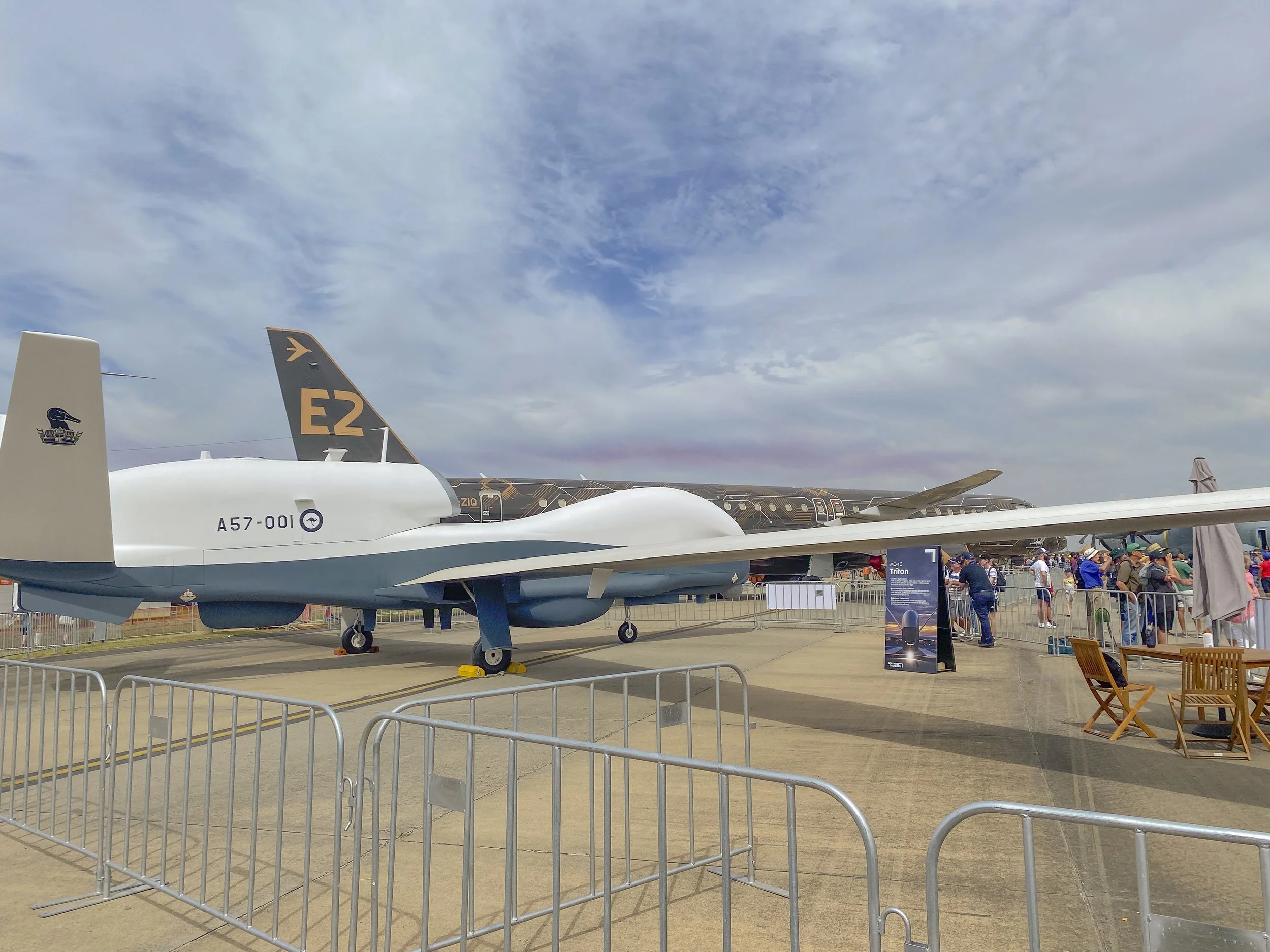 An aircraft on display at an airshow with spectators walking around. The aircraft has a white and gray body, with a desert camouflage jet in the background.