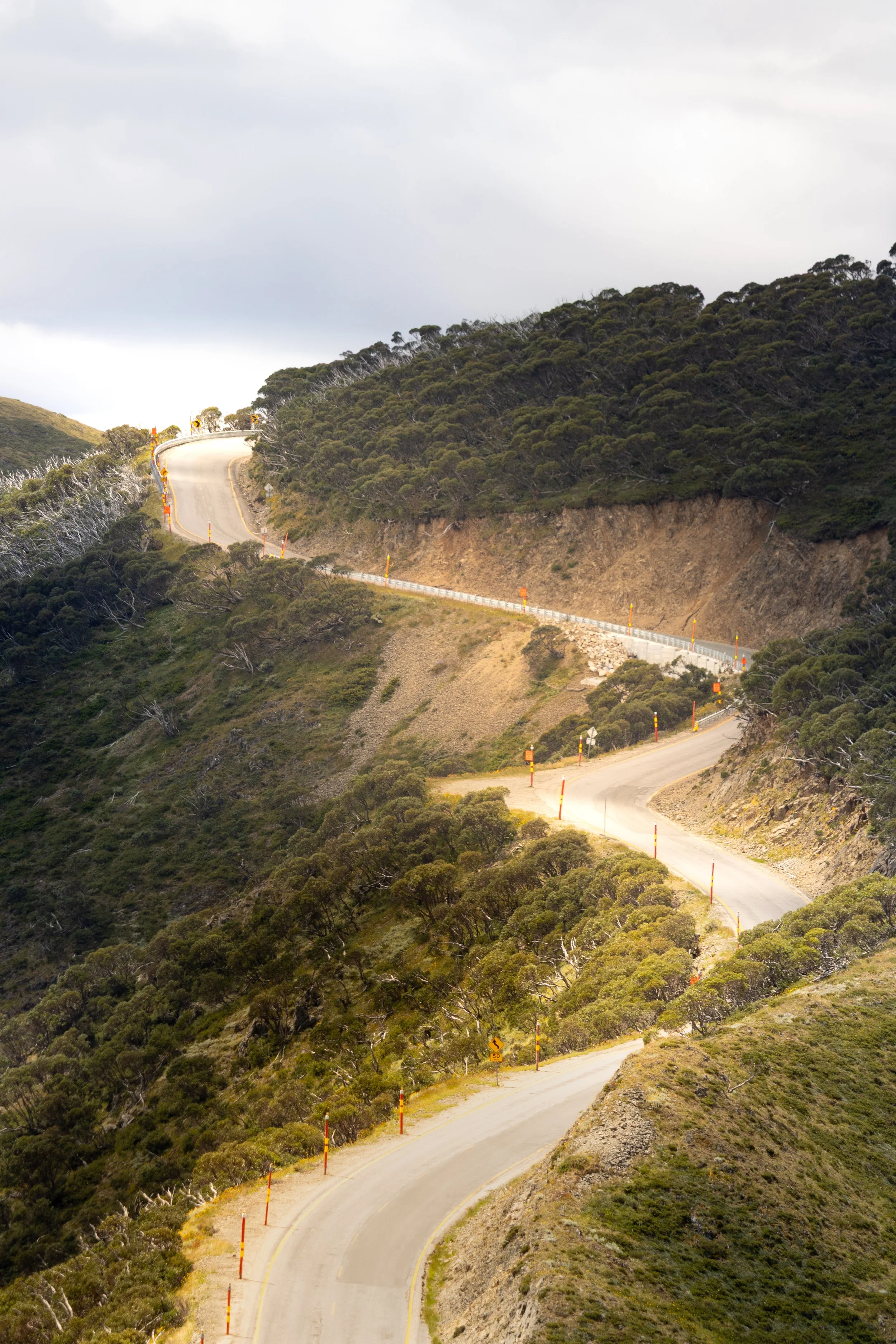 Winding mountain road with orange and yellow safety markers, surrounded by green shrubbery and hills under a cloudy sky.