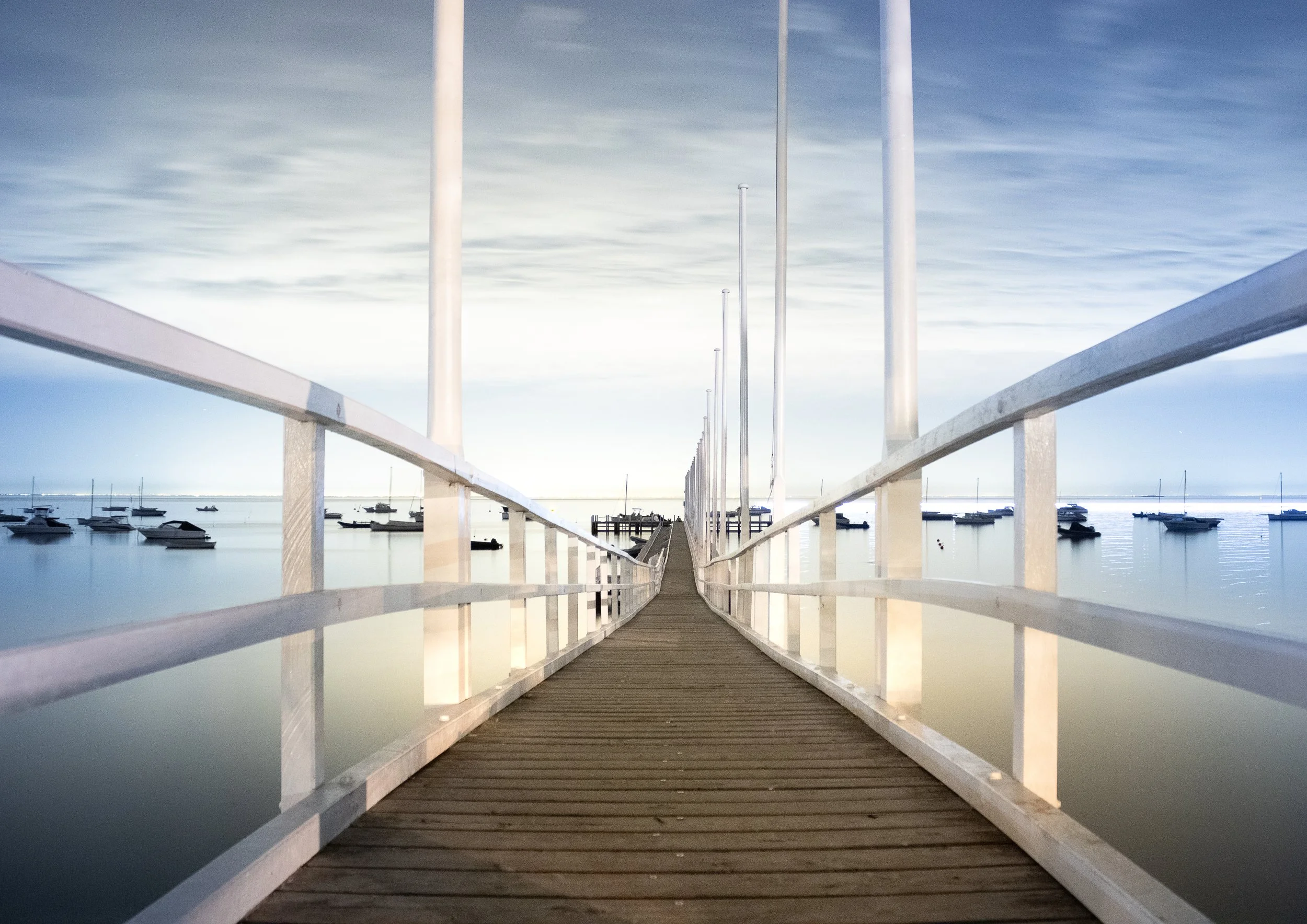 A wooden pier extends into a calm body of water with boats anchored on either side. The sky is overcast, and the scene is tranquil.