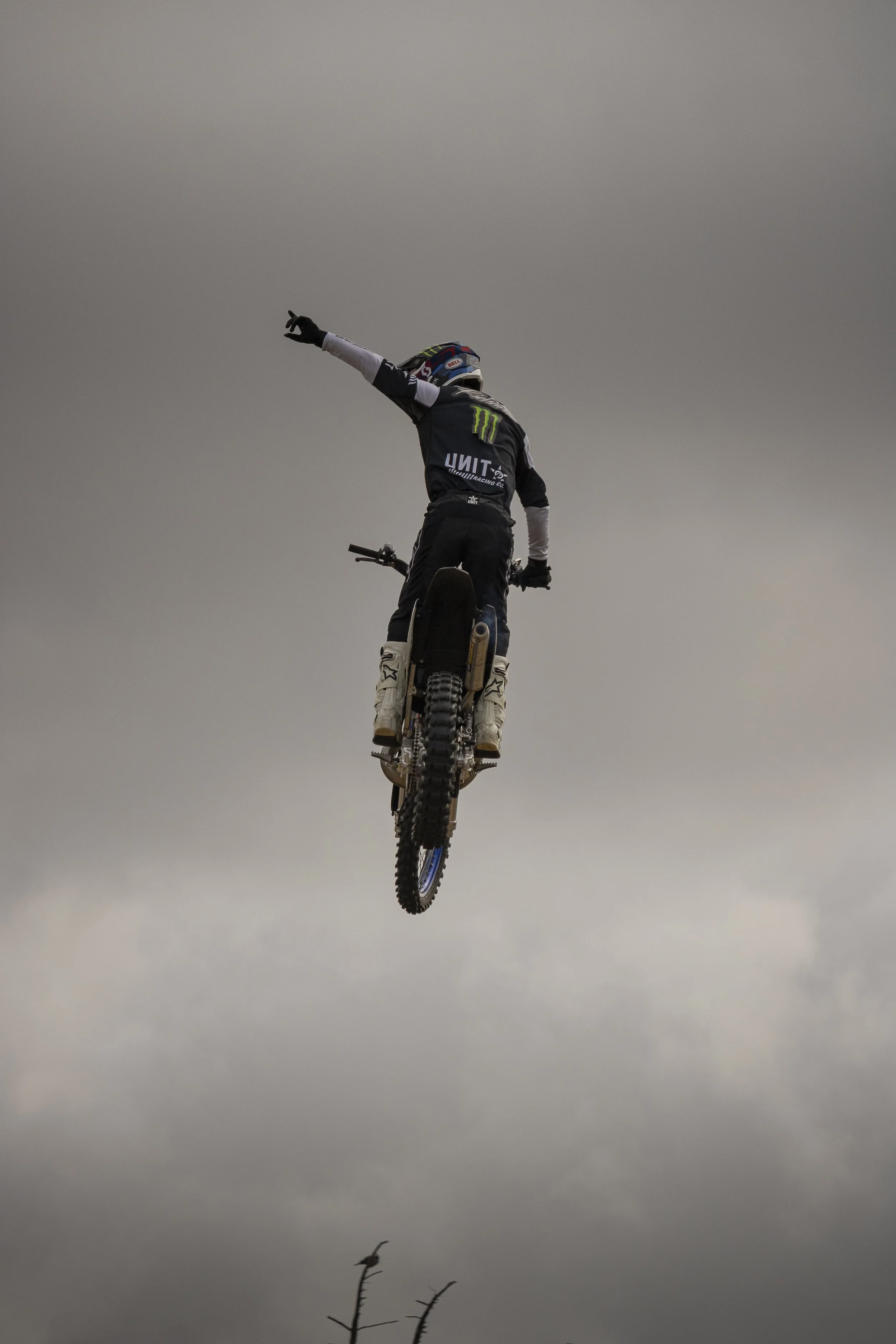 Motorcycle stunt rider airborne against cloudy sky, pointing with left arm extended.