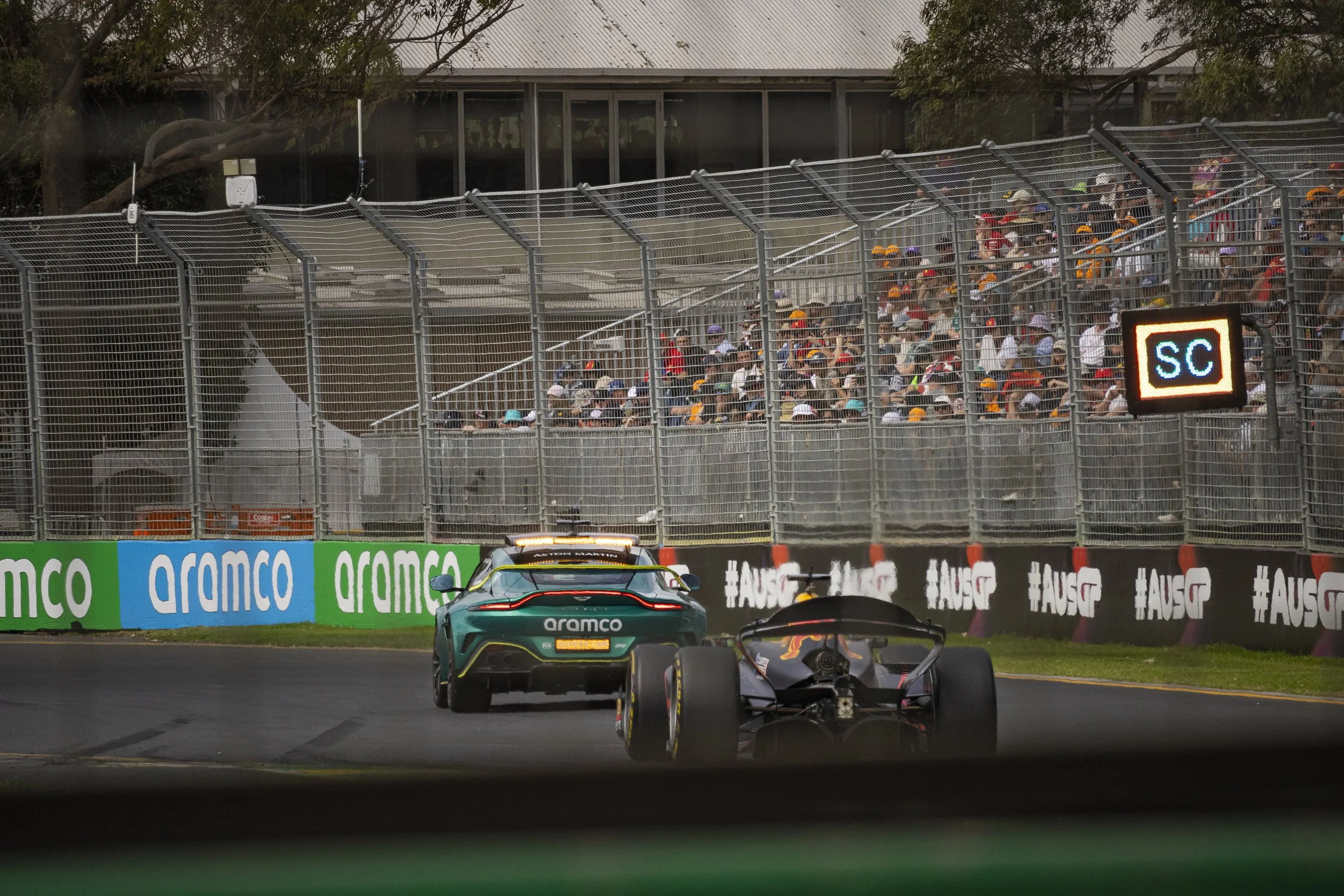 A race car on a track with a green and black design and a Formula 1 style, passing another open-wheel race car with a black and orange design. Spectators in colorful hats are visible behind a tall, metal fence.