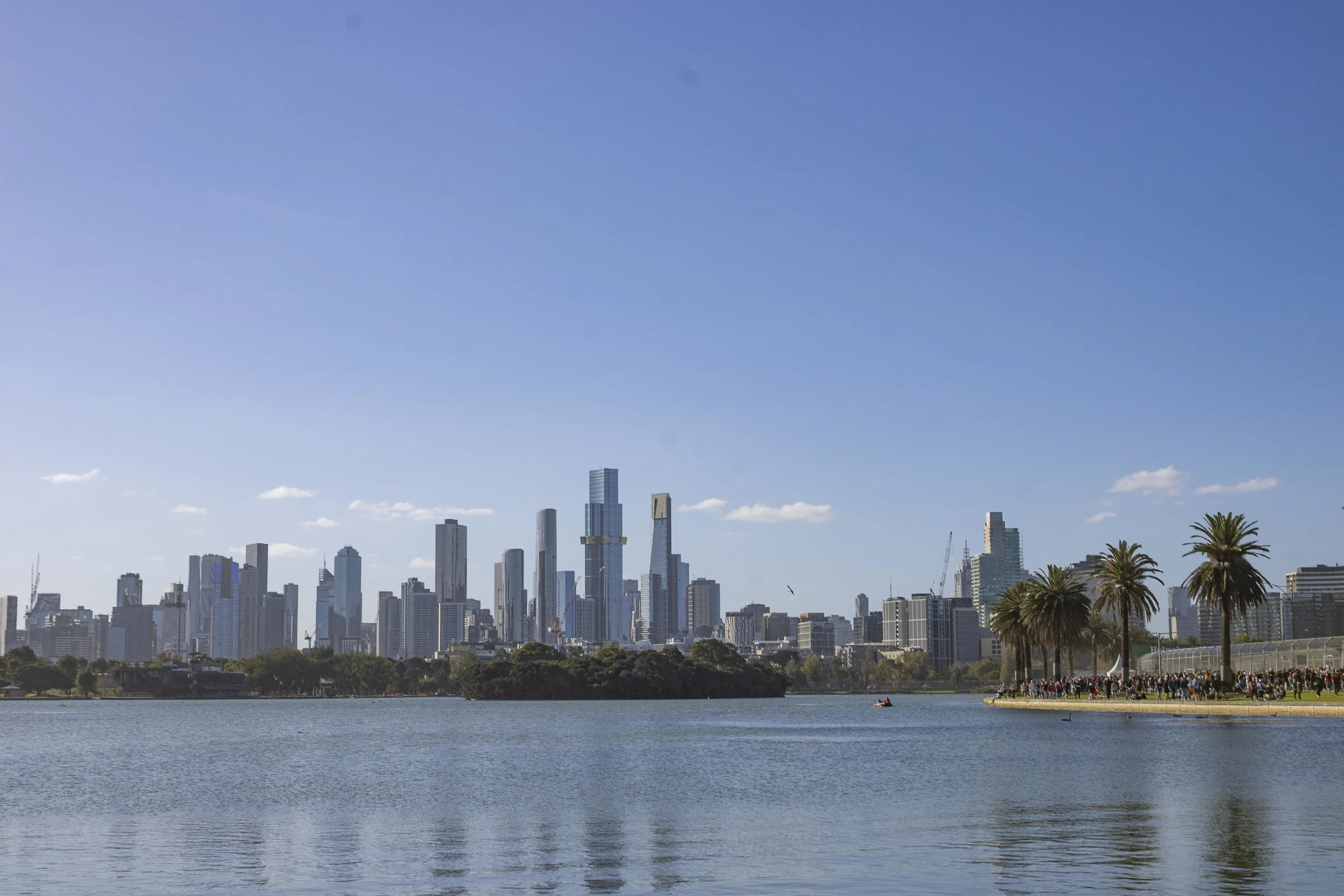 City skyline with tall buildings and skyscrapers across a body of water, with palm trees and a crowd of people on the right side of the shoreline.