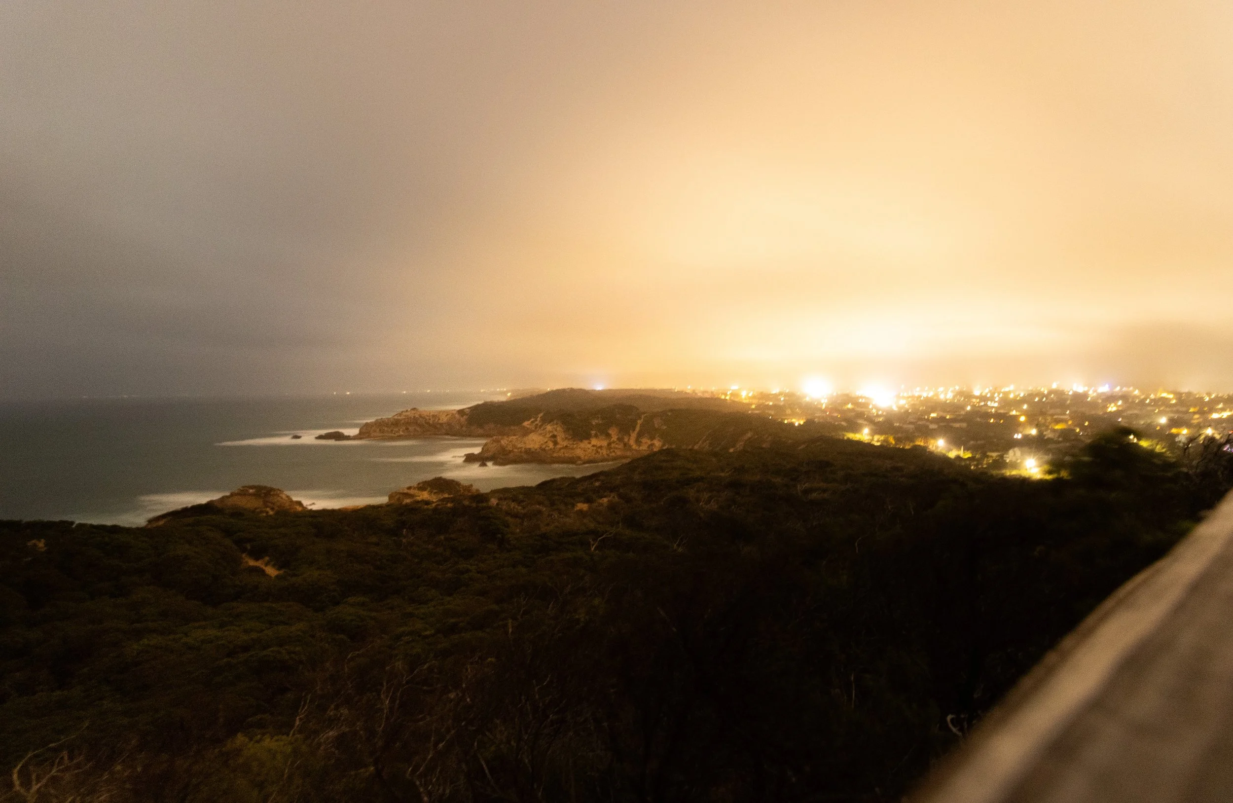 Nighttime view of the coastline with waves crashing against rocky cliffs, illuminated by city lights in the distance under a cloudy sky.