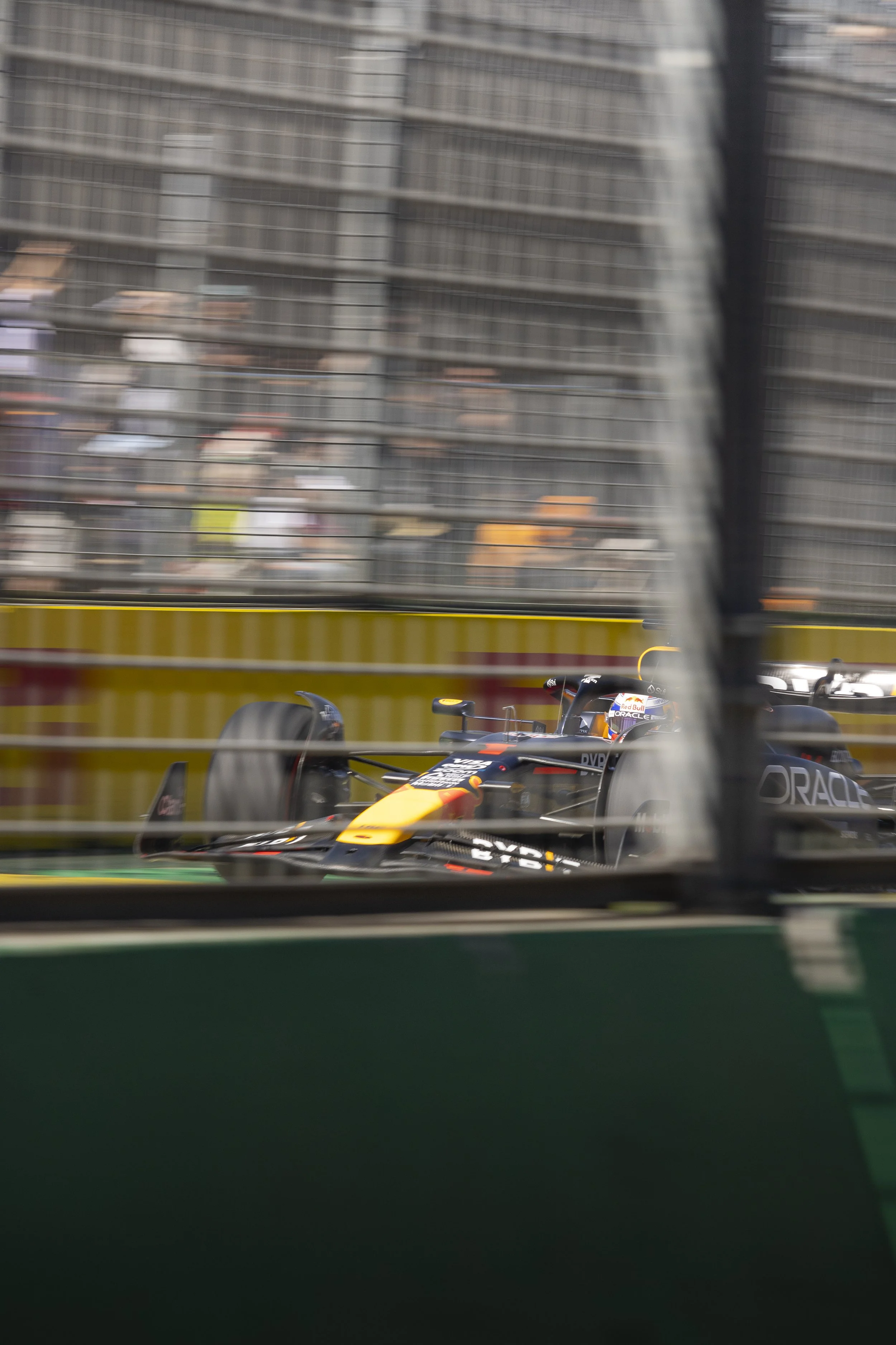 Race car speeding on a track, seen through a fence with a background of tall buildings.