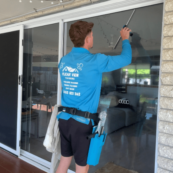 A window cleaner using a squeegee to clean a large glass window on a house. The worker is wearing a blue shirt with company branding and has cleaning tools hanging from his waist.