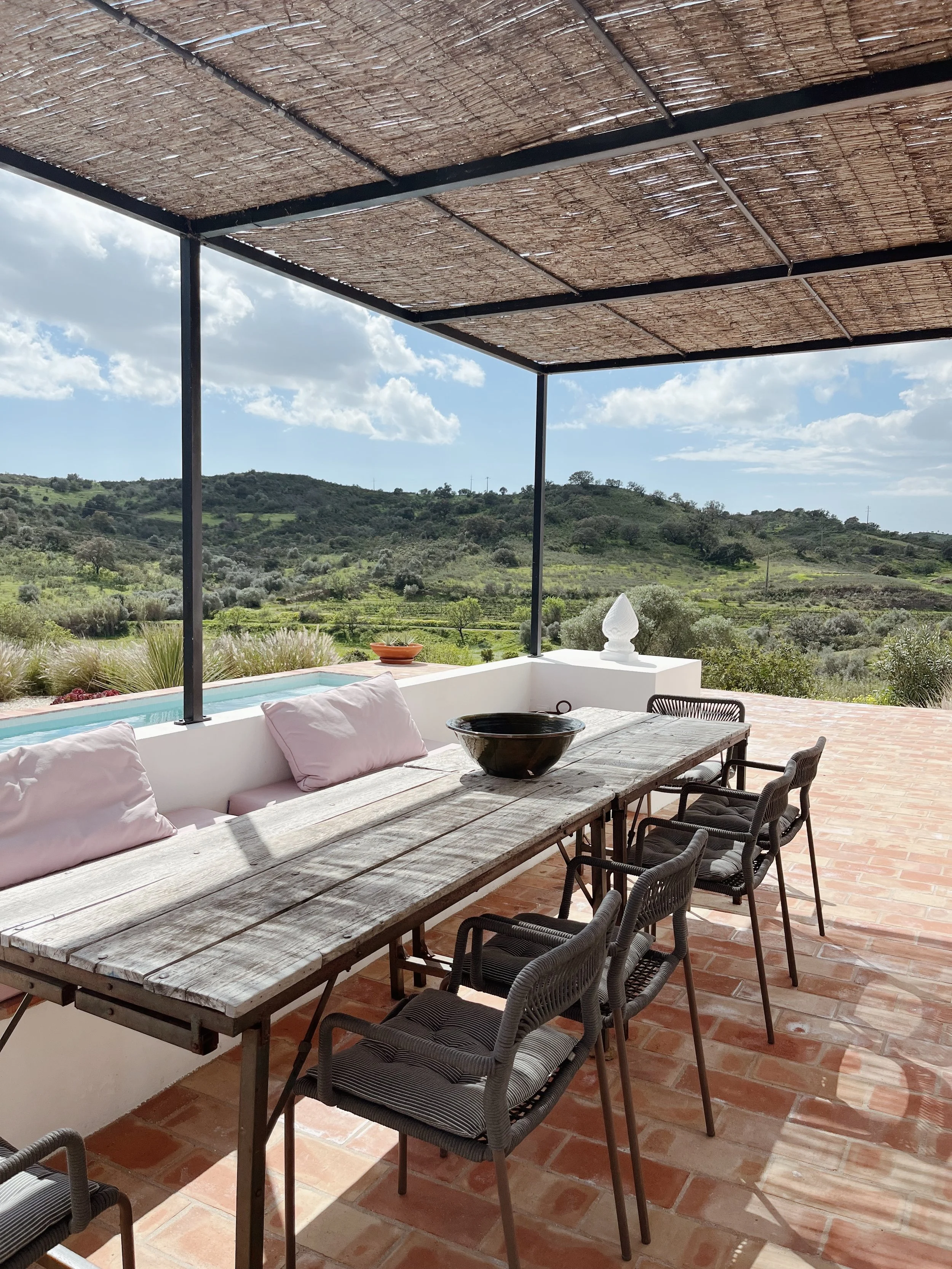 Balcon avec un long table en bois, six chaises en osier avec coussins, et des coussins blancs sur le banc en pierre. Vue sur une campagne verdoyante avec des collines et des arbres, sous un ciel partiellement nuageux.