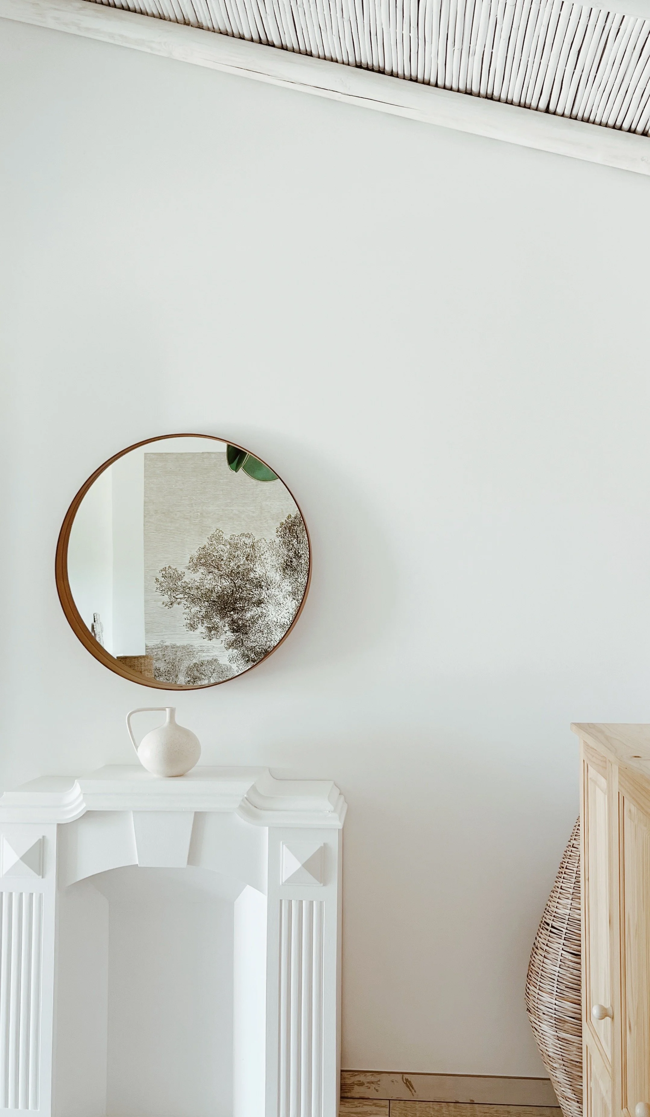 Un mur blanc avec un miroir rond au centre, une cheminée blanche en dessous portant un vase blanc, et un meuble en bois clair à droite avec un panier en osier.