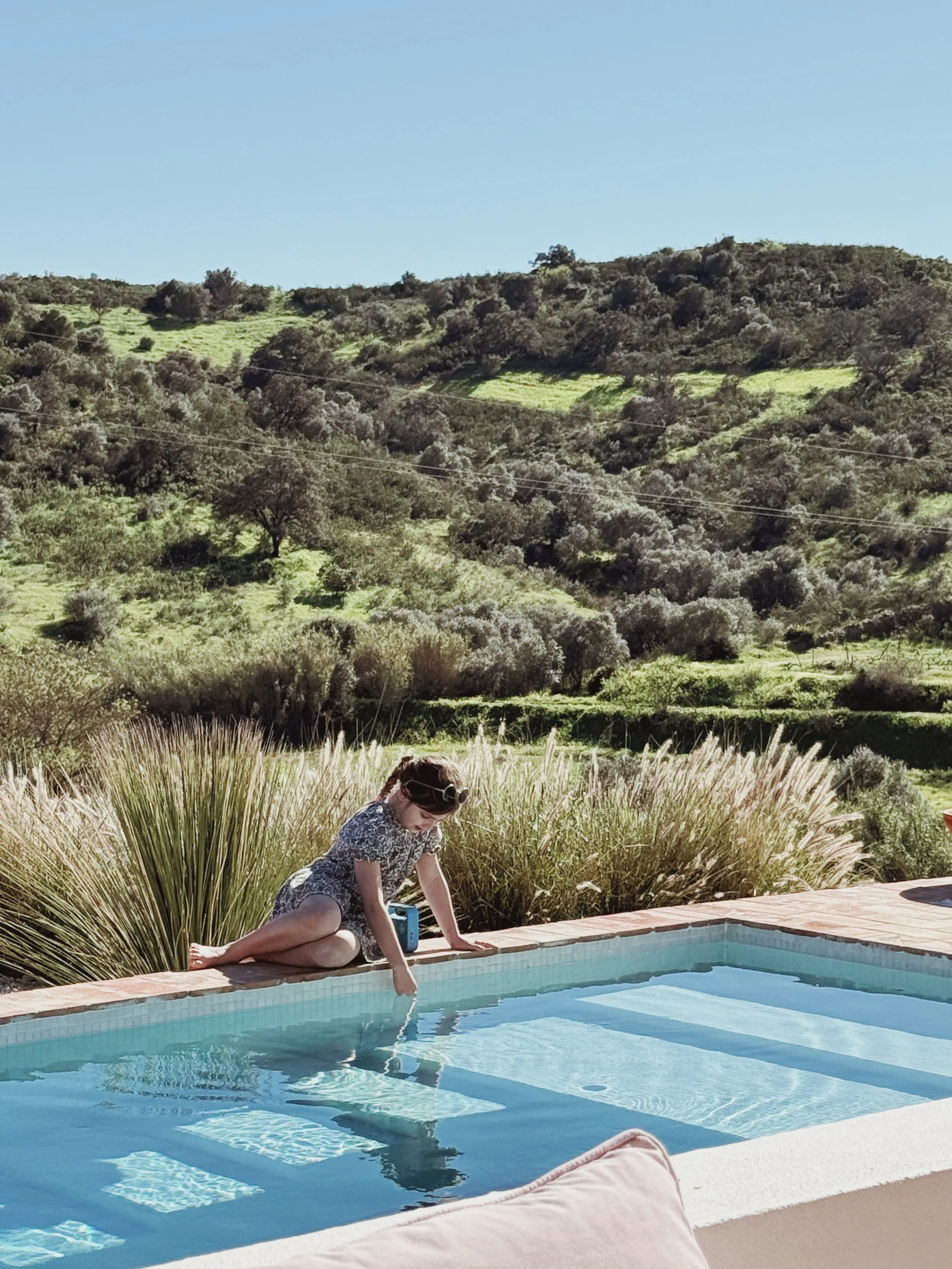 Une jeune fille en robe à fleurs, assise au bord d'une piscine, touche l'eau avec son doigt, avec un paysage de collines verdoyantes et arbres en arrière-plan.