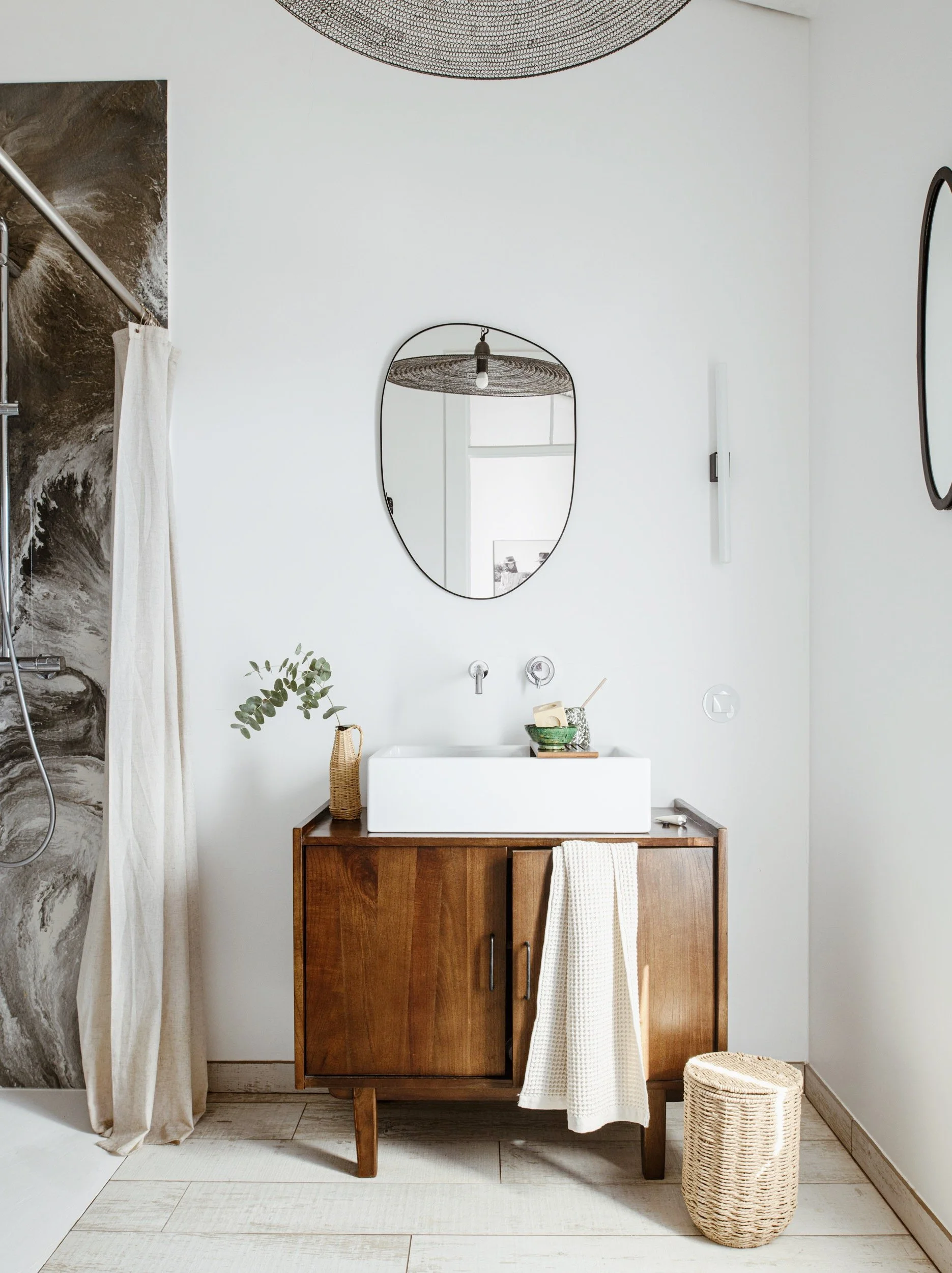 Salle de bain avec un meuble en bois, une vasque blanche et un miroir mural, décoration minimaliste, éléments naturels comme une plante et un panier en osier.