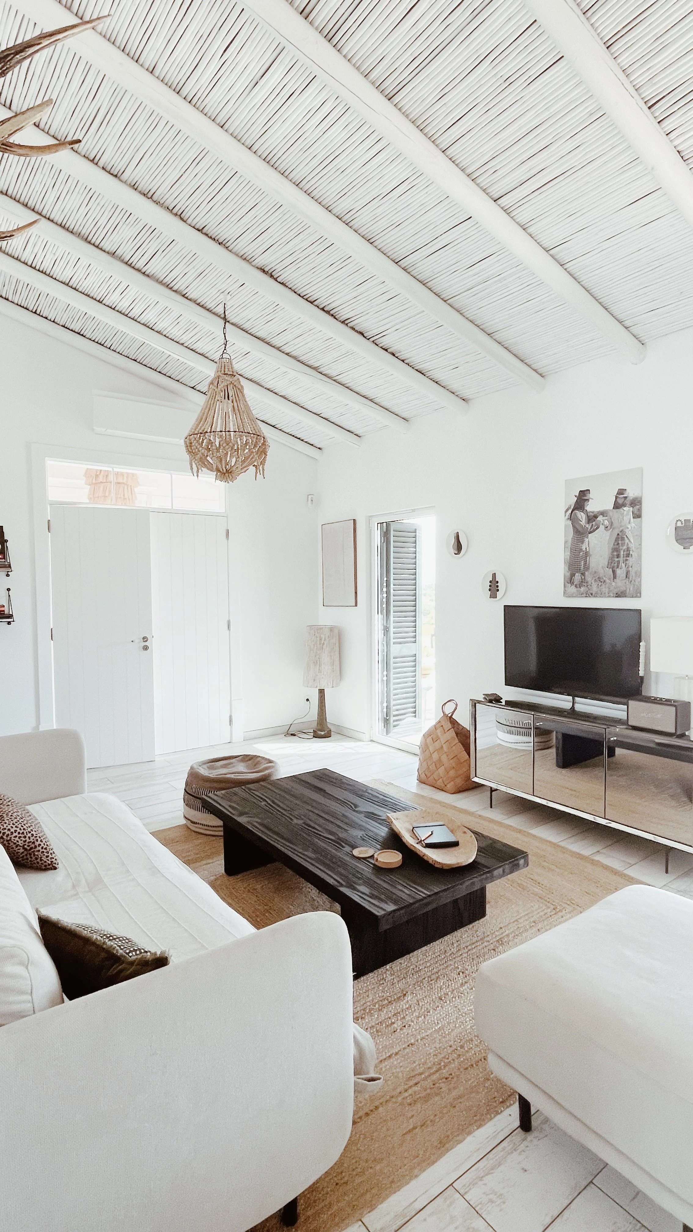 Salon lumineux avec canapé blanc, table basse en bois foncé, TV, lampadaire, et décorations murales. Plafond en bois clair untouché, porte-fenêtre en verre ouvrant vers l'extérieur.