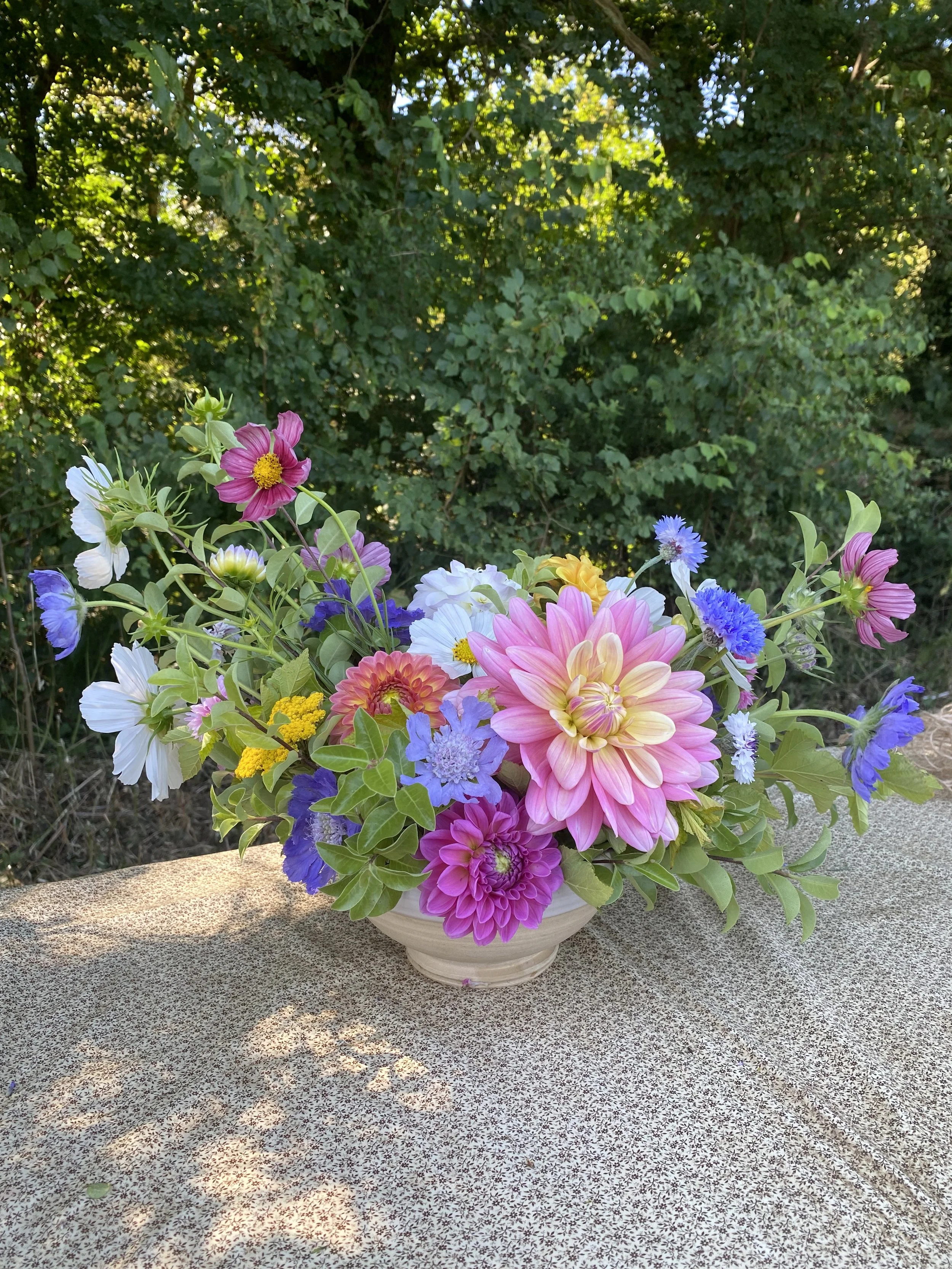 Un vase rempli de fleurs colorées, y compris des dahlias, fleurs de cosmos, et autres, posé sur une table-texture avec un fond de végétation verte dense.