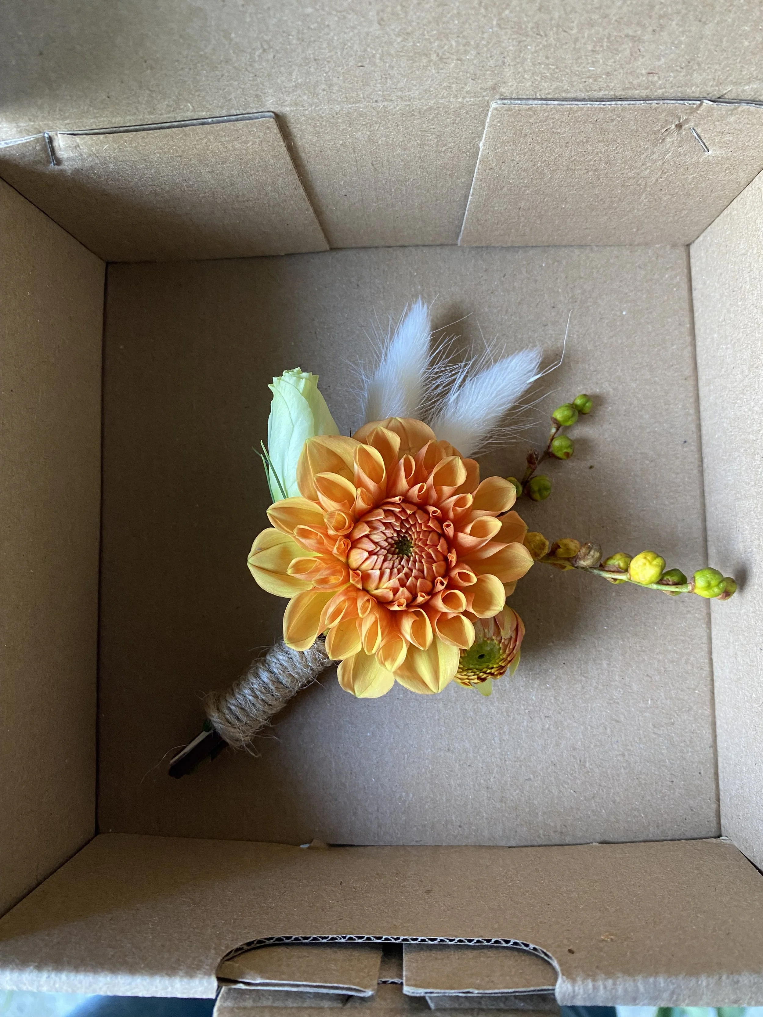 Un bouquet de fleurs y compris une grosse fleur orange, quelques petites fleurs jaunes, des bourgeons verts, et des plumes blanches, toutes placées dans une boîte en carton