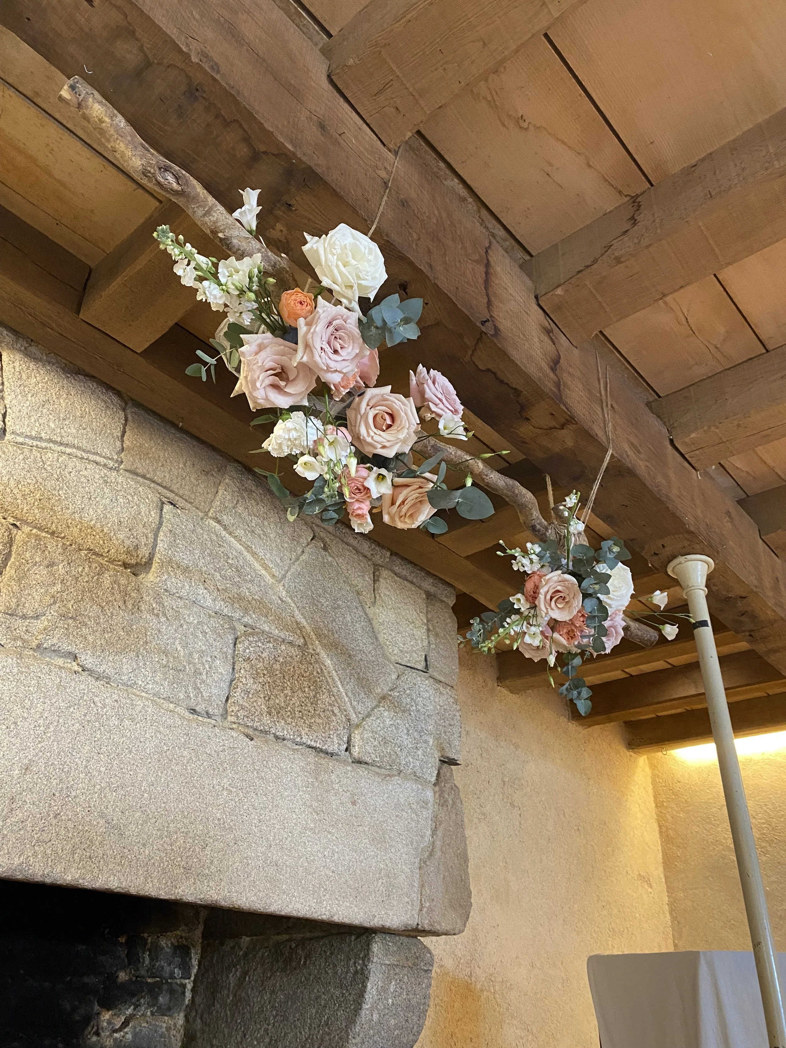 Une décoration florale composée de roses roses, blanches et d'autres fleurs, suspendue sous un plafond en bois au-dessus d'une pierre murale.