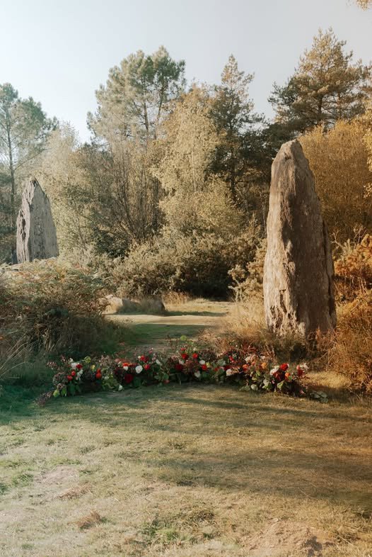 Un site de cérémonie avec deux grands menhirs, des fleurs en bouquets posés au sol, entouré d'arbres et de buissons dans un cadre naturel.